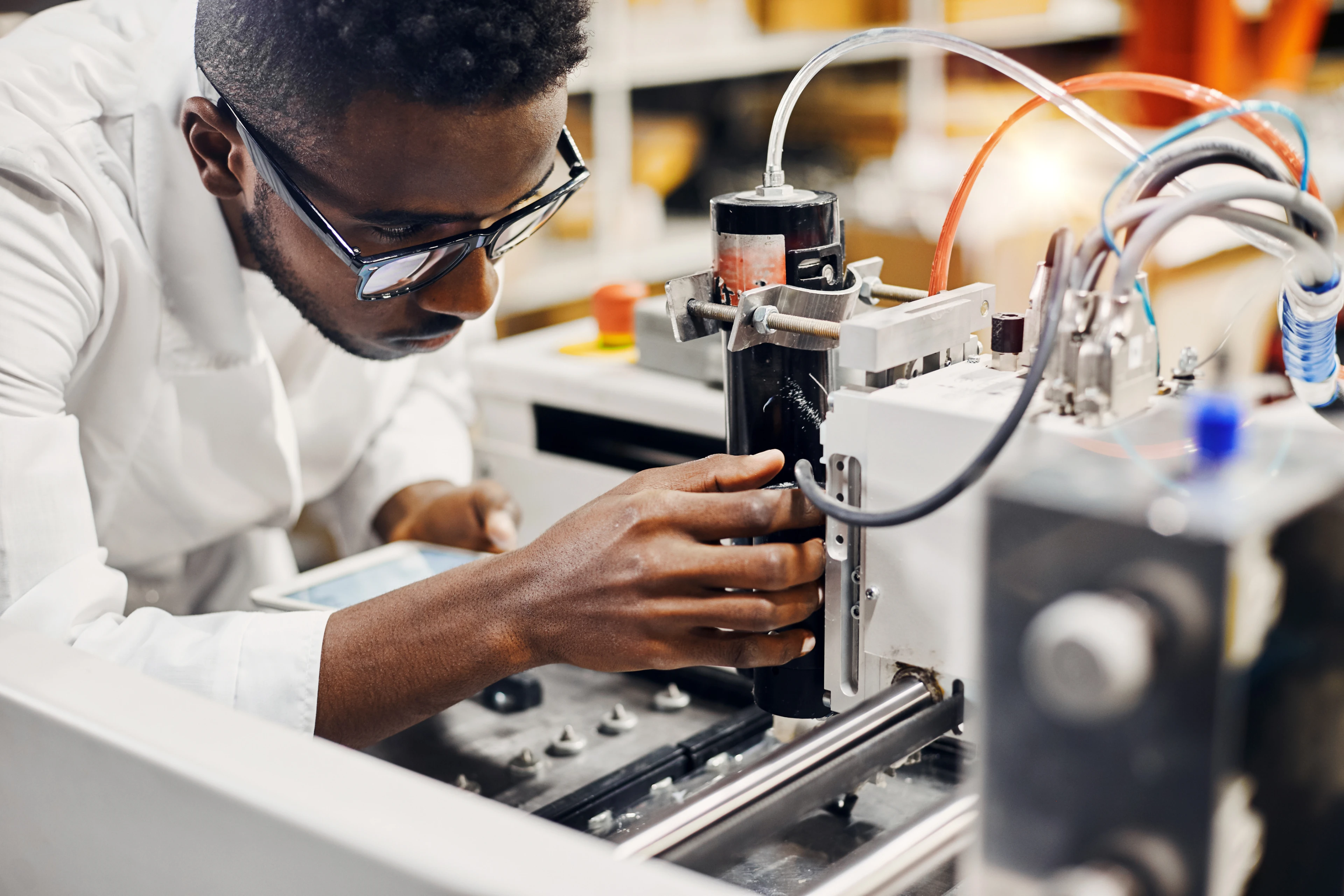 A lab technician in safety glasses works with equipment while holding a tablet, representing a regulated UK assessment completed through an online proctoring solution.