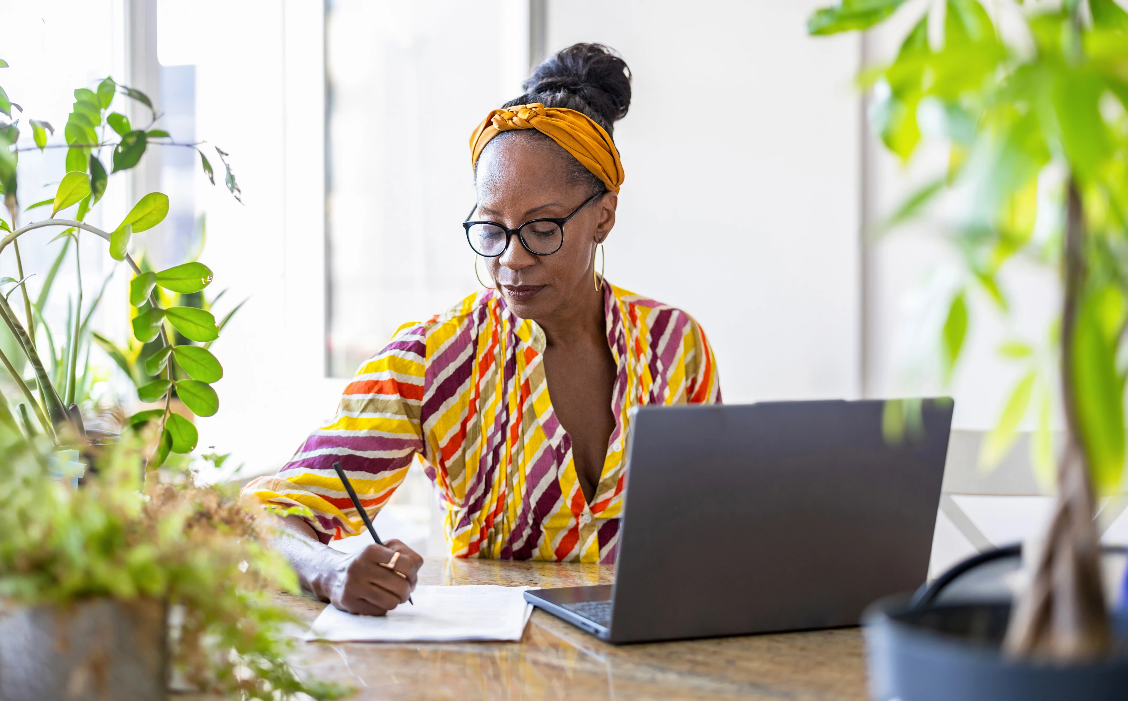A woman writes notes at a desk with a laptop, surrounded by plants, representing a condominium manager completing licensing coursework online.