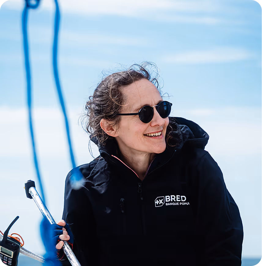 Femme souriante portant des lunettes de soleil et une veste BRED Banque Populaire sur un bateau par temps clair.