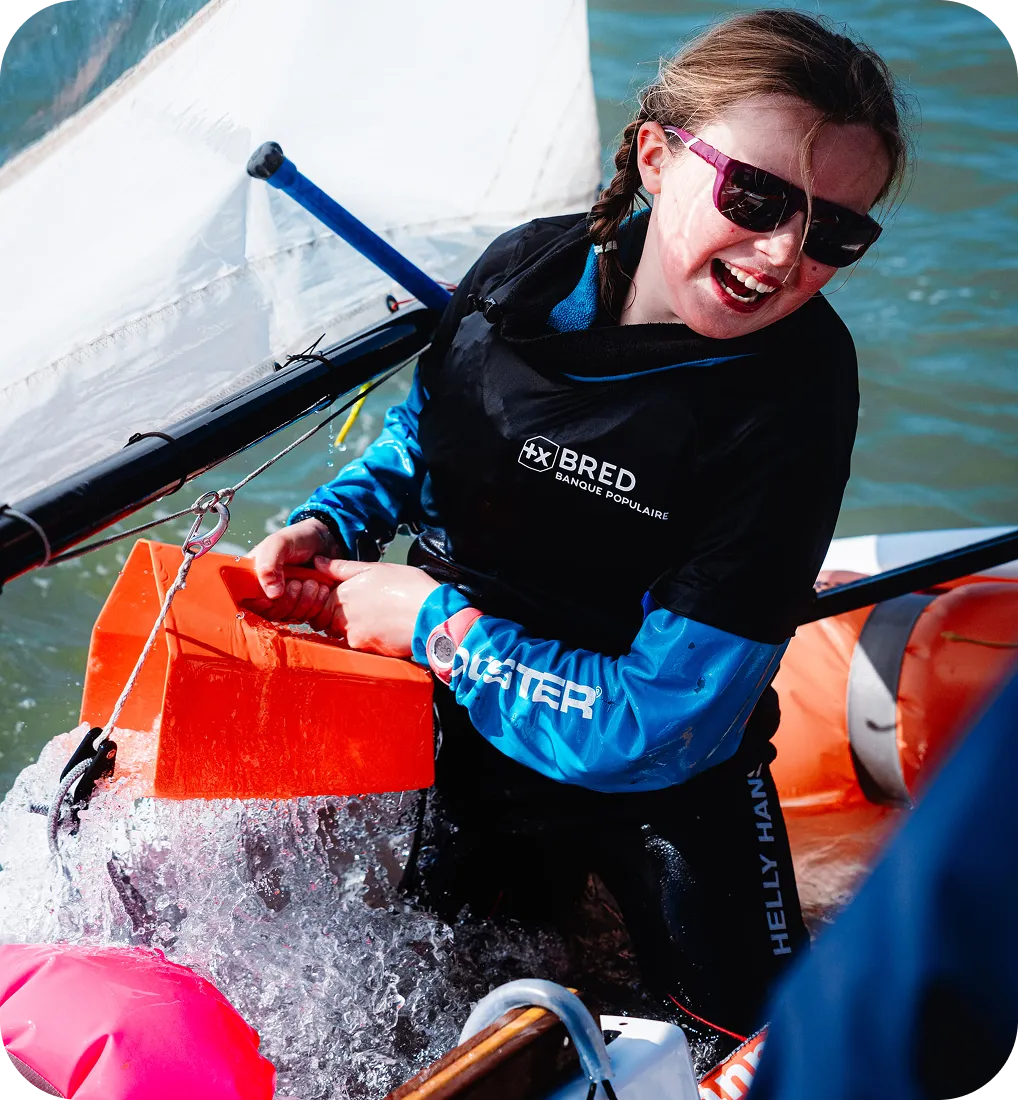 Jeune fille souriante en tenue de voile récupérant de l'eau dans un bateau avec un seau orange.