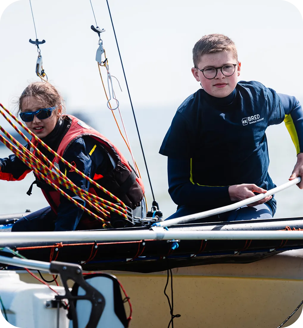 Deux enfants portant des lunettes et des gilets de sauvetage naviguent sur un petit voilier avec des cordes colorées.