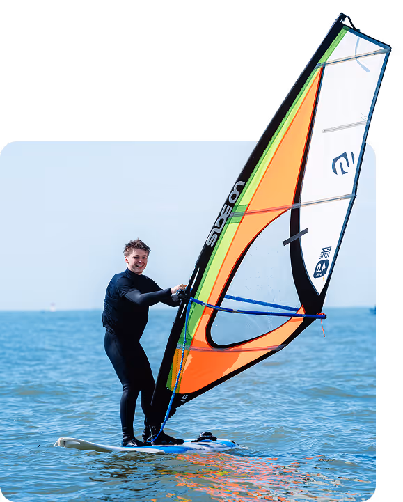 Un jeune homme en combinaison de plongée tient une grande voile orange, verte et blanche sur une planche de planche à voile dans l'eau.