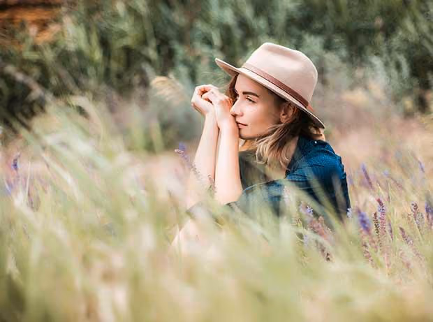 Frau mit Hut sitzt nachdenklich in einem Feld mit hohen Gräsern und lila Wildblumen.