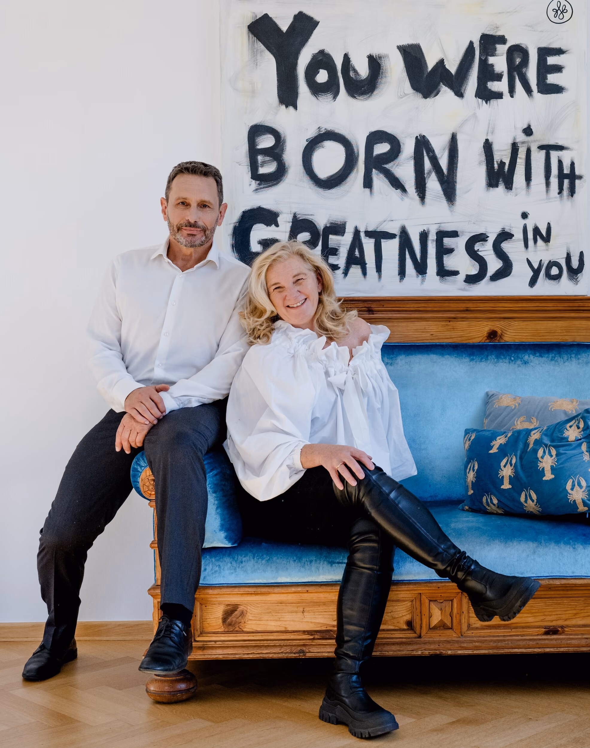 Ein Mann und eine Frau sitzen lächelnd auf einem blauen Sofa vor einem Bild mit dem Text ‚You were born with greatness‘.