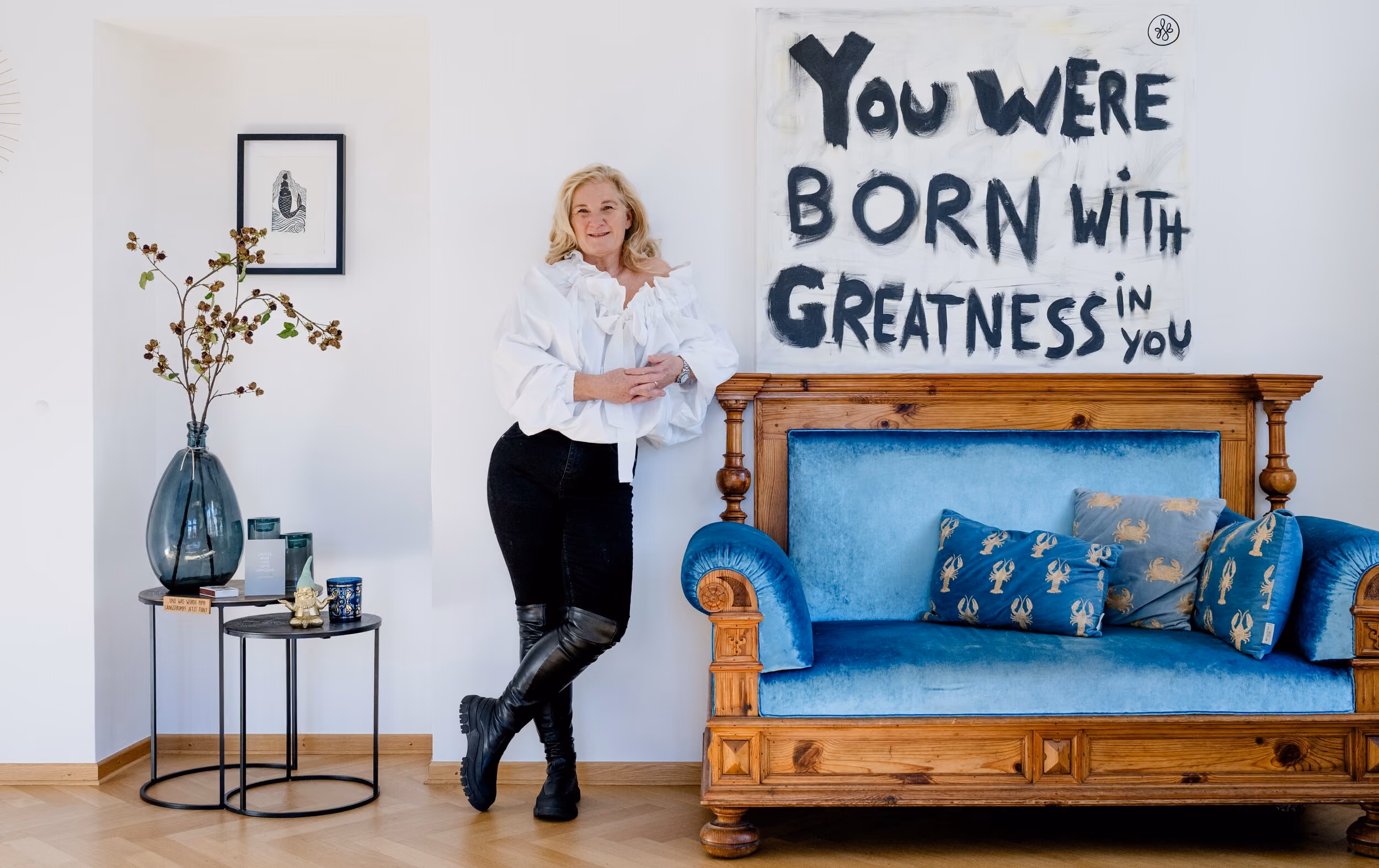 Frau lehnt an weißer Wand neben blauem Sofa mit Holzrahmen und motivierendem Wandbild.