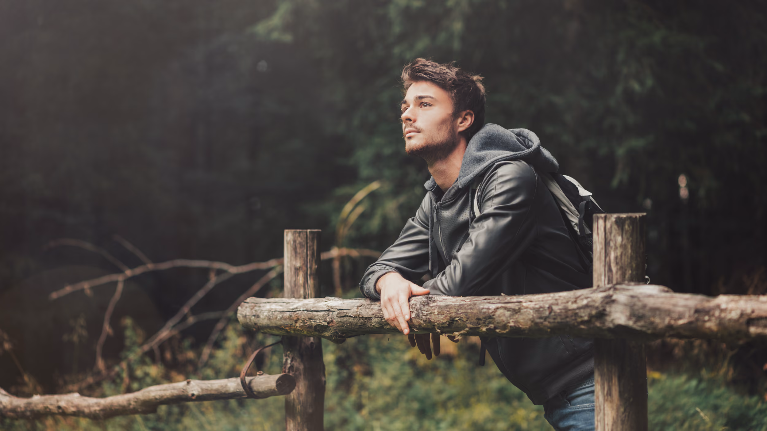 Junger Mann lehnt an einem Holzzaun und blickt nachdenklich in die Ferne im Wald.