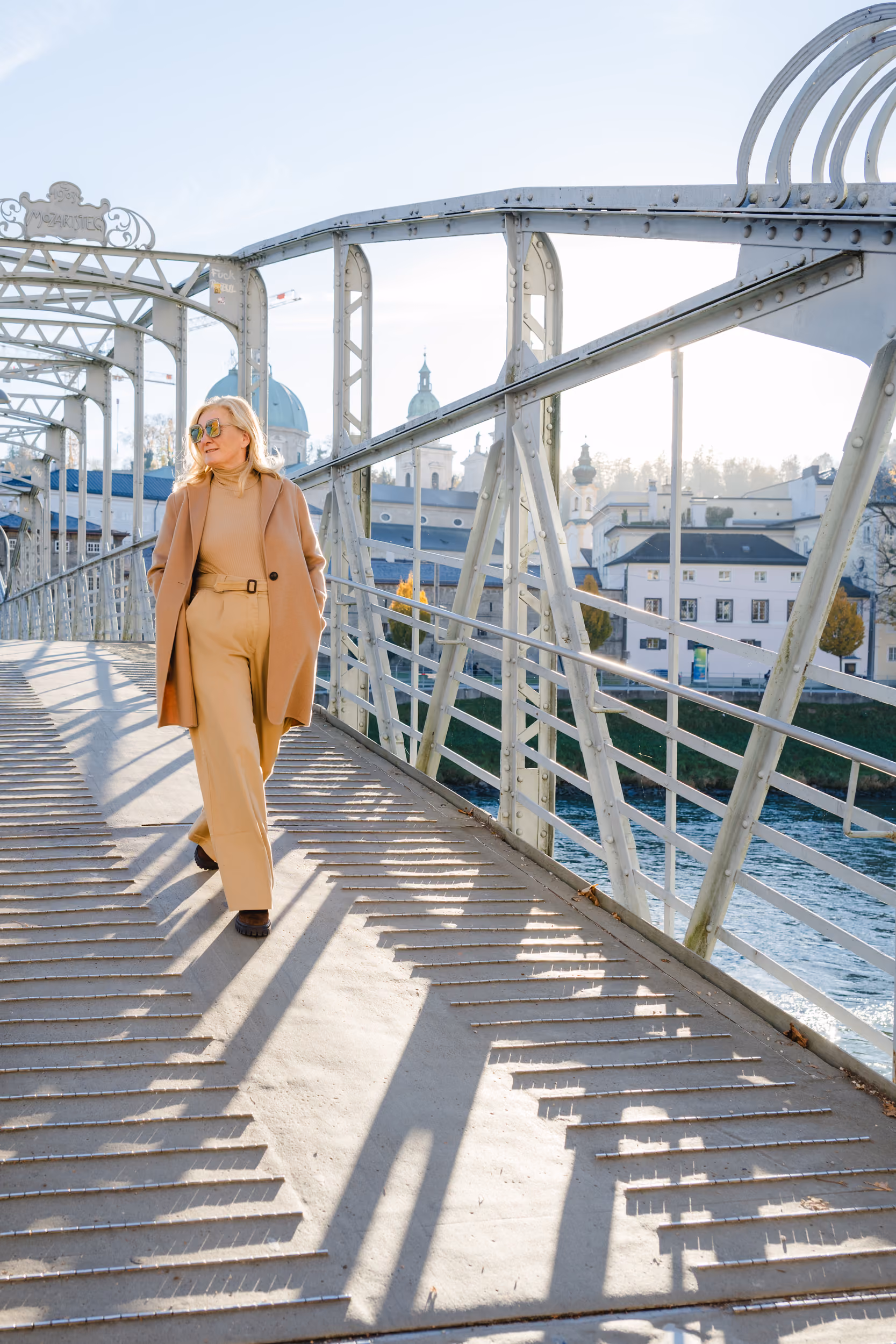 Frau in beigem Mantel und Hose geht auf einer Metallbrücke mit Gebäuden und Fluss im Hintergrund.