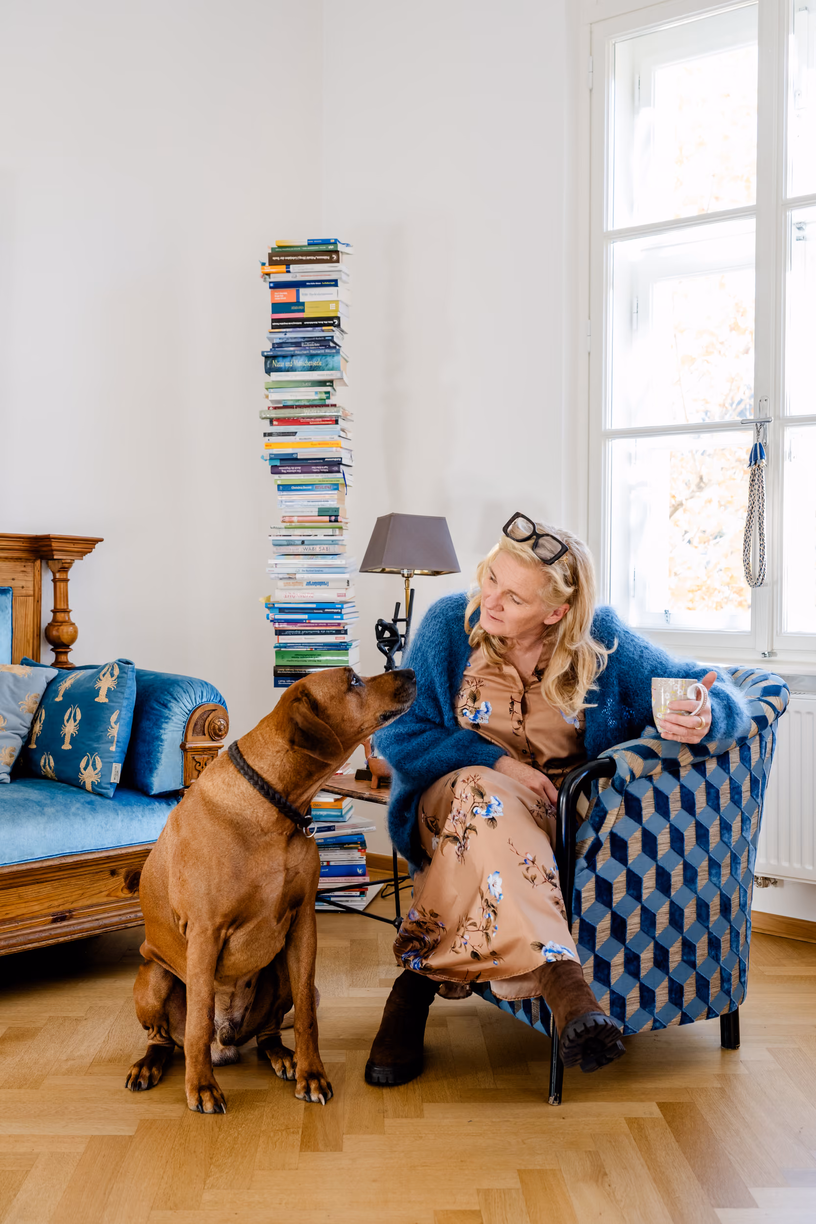 Frau sitzt in Sessel mit Tasse und sieht großen Hund an, neben Bücherstapel und Sofa im Wohnzimmer.