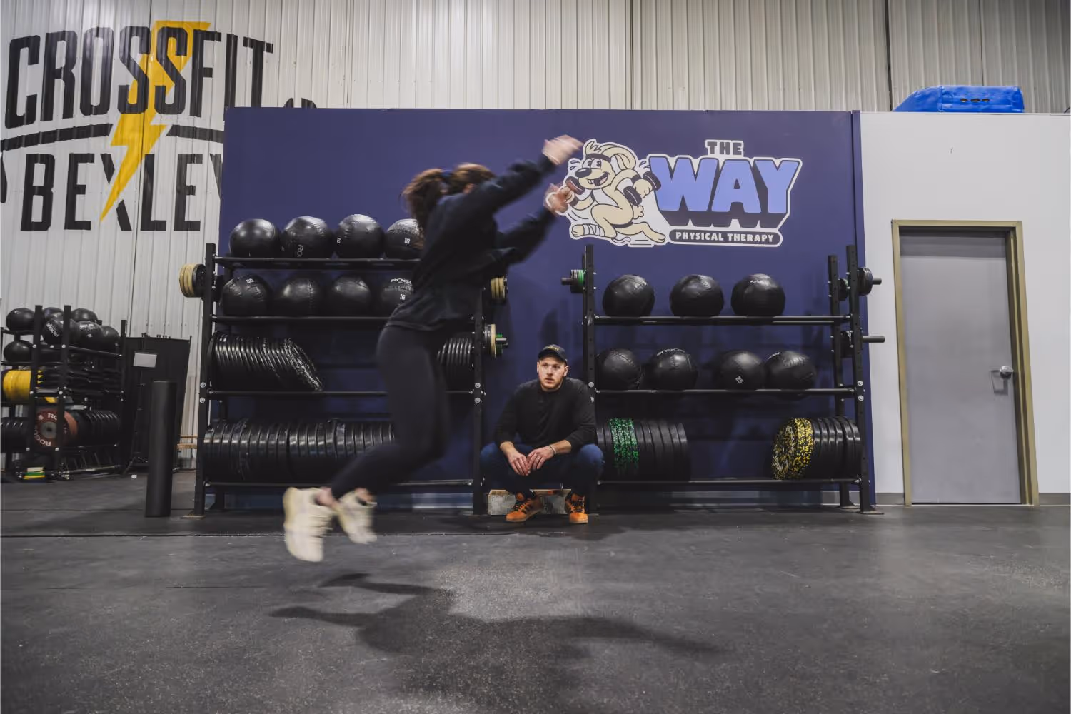 A woman performing a jumping exercise in a CrossFit gym with a man squatting and watching behind her.