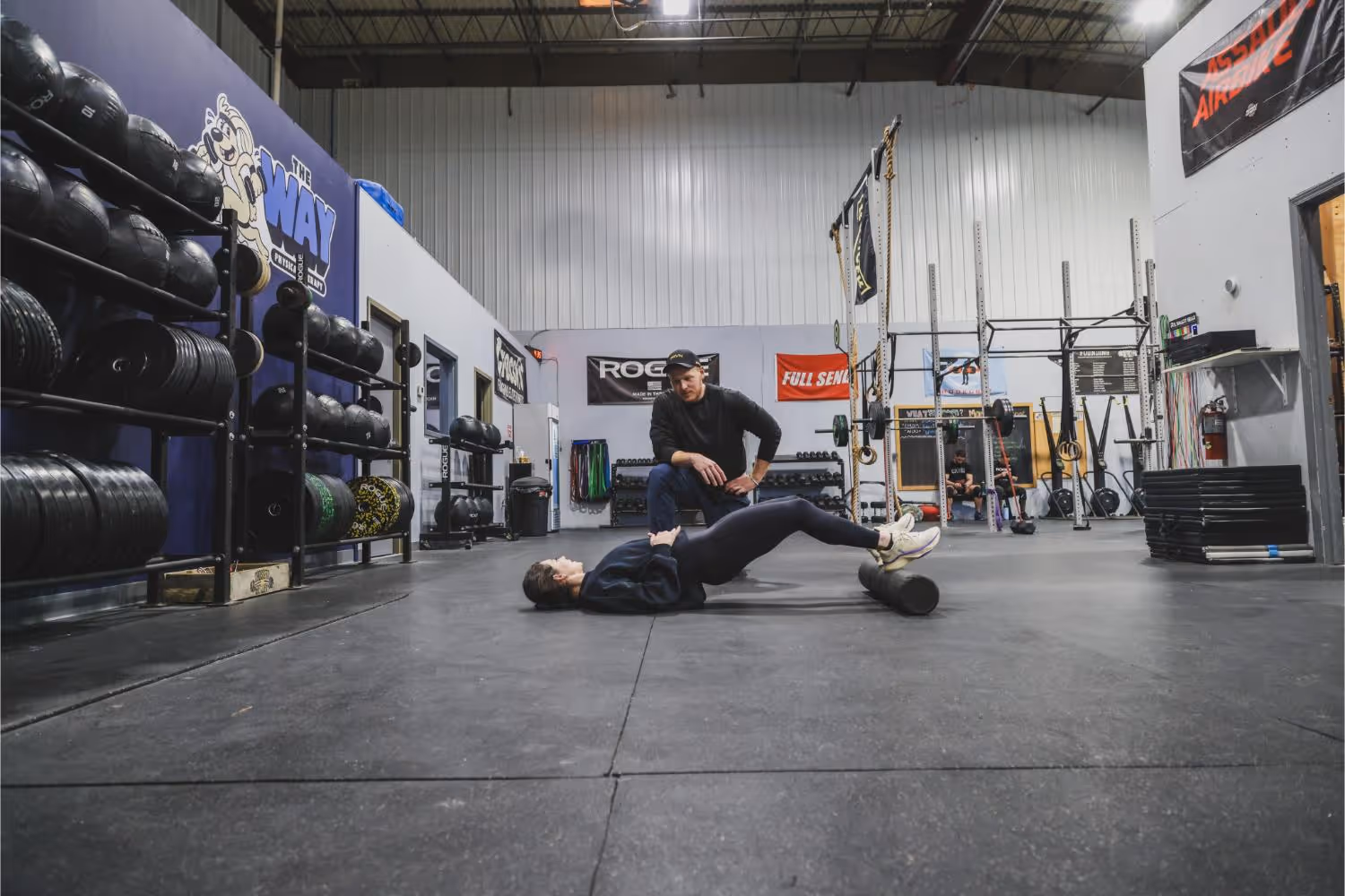 A man in black athletic wear is using a foam roller on his calves while lying on the gym floor, with a trainer kneeling and instructing him in a spacious gym with weights and exercise equipment.