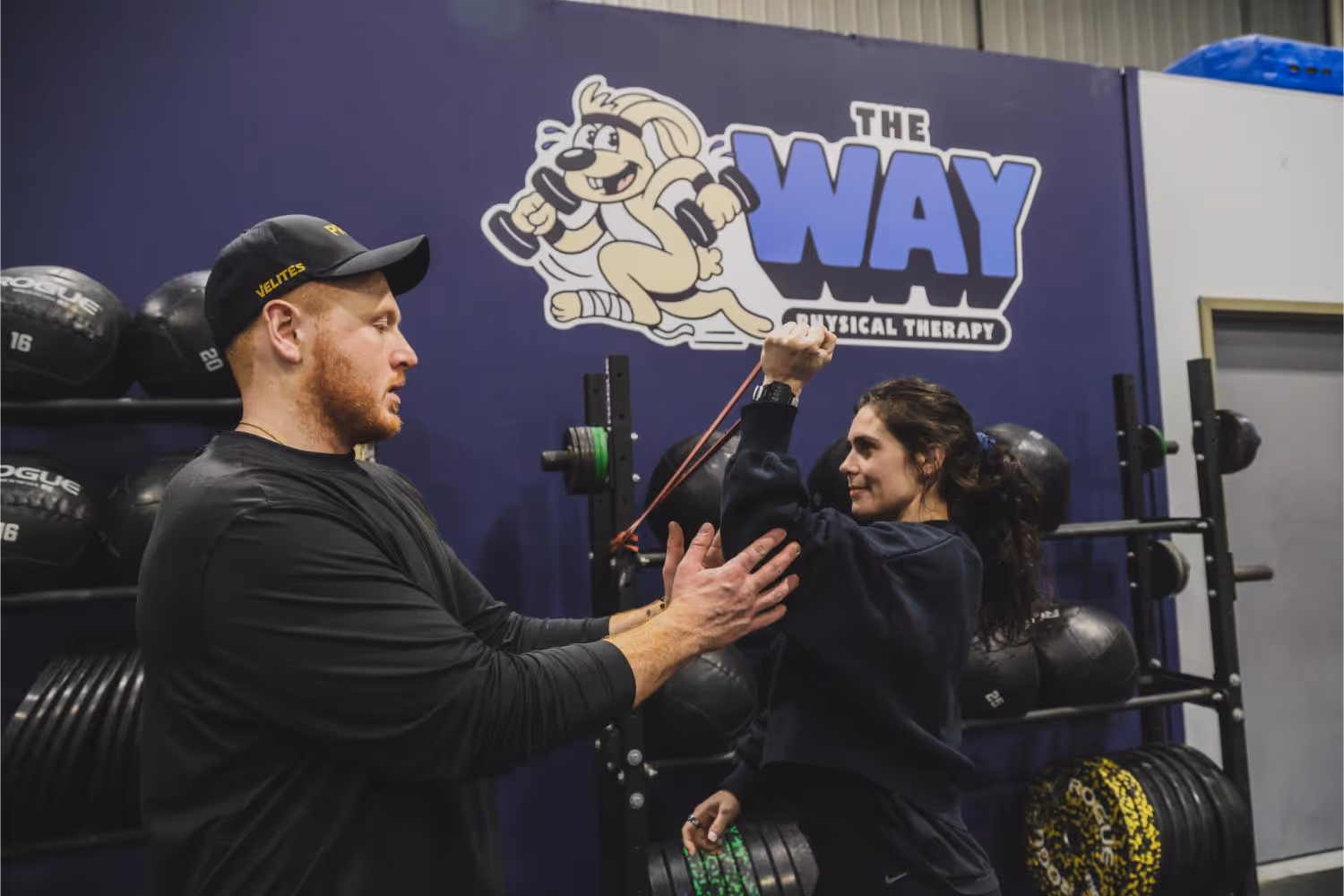 Physical therapist assisting a woman with resistance band arm exercise in a gym with weights and a 'The Way Physical Therapy' sign on the wall.
