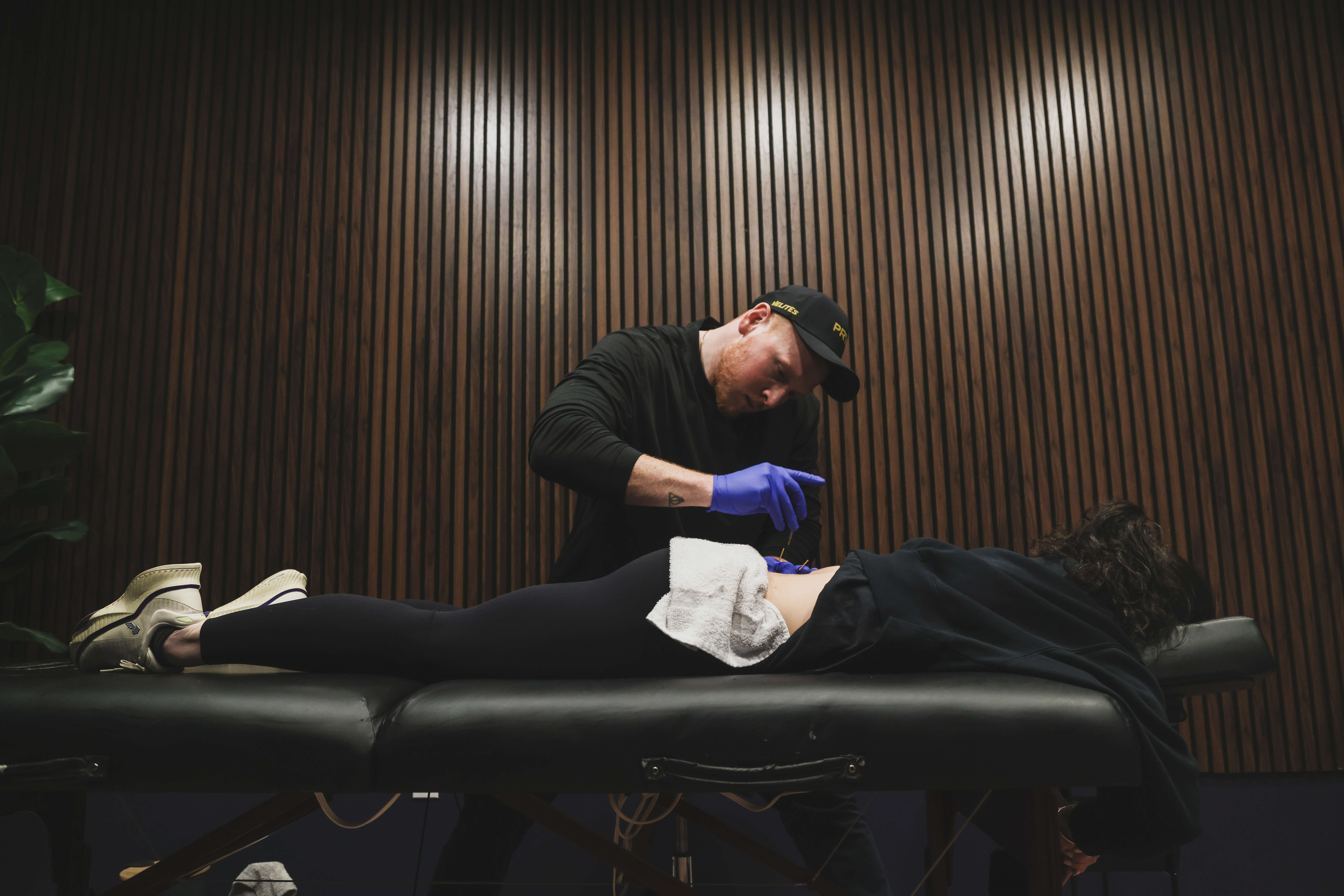 A tattoo artist wearing gloves and a cap is tattooing a person's lower back while they lie face down on a black padded table.