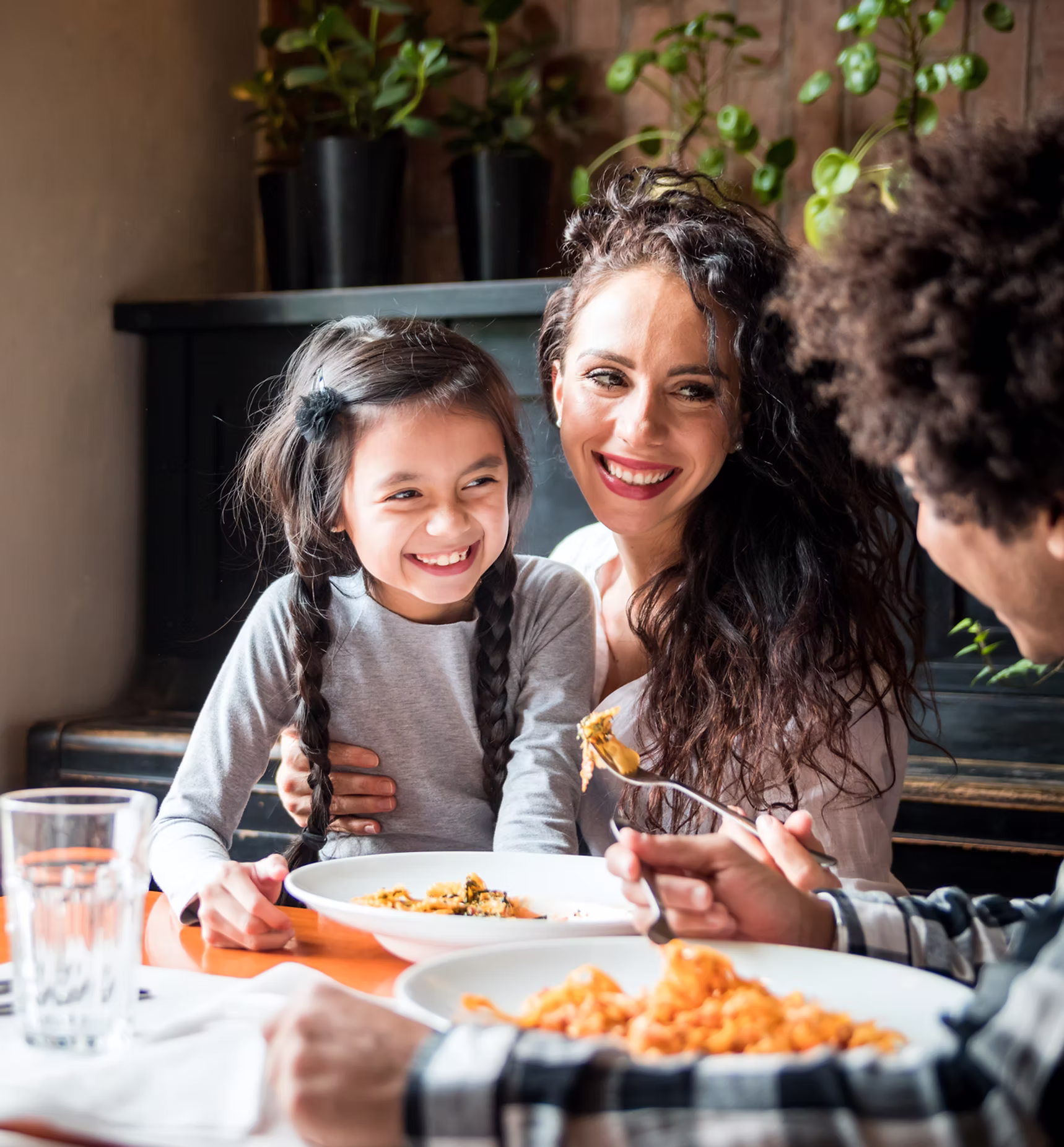 Smiling woman and young girl with braided hair sitting together at a table with plates of pasta, sharing a joyful moment while a man offers food with a fork.