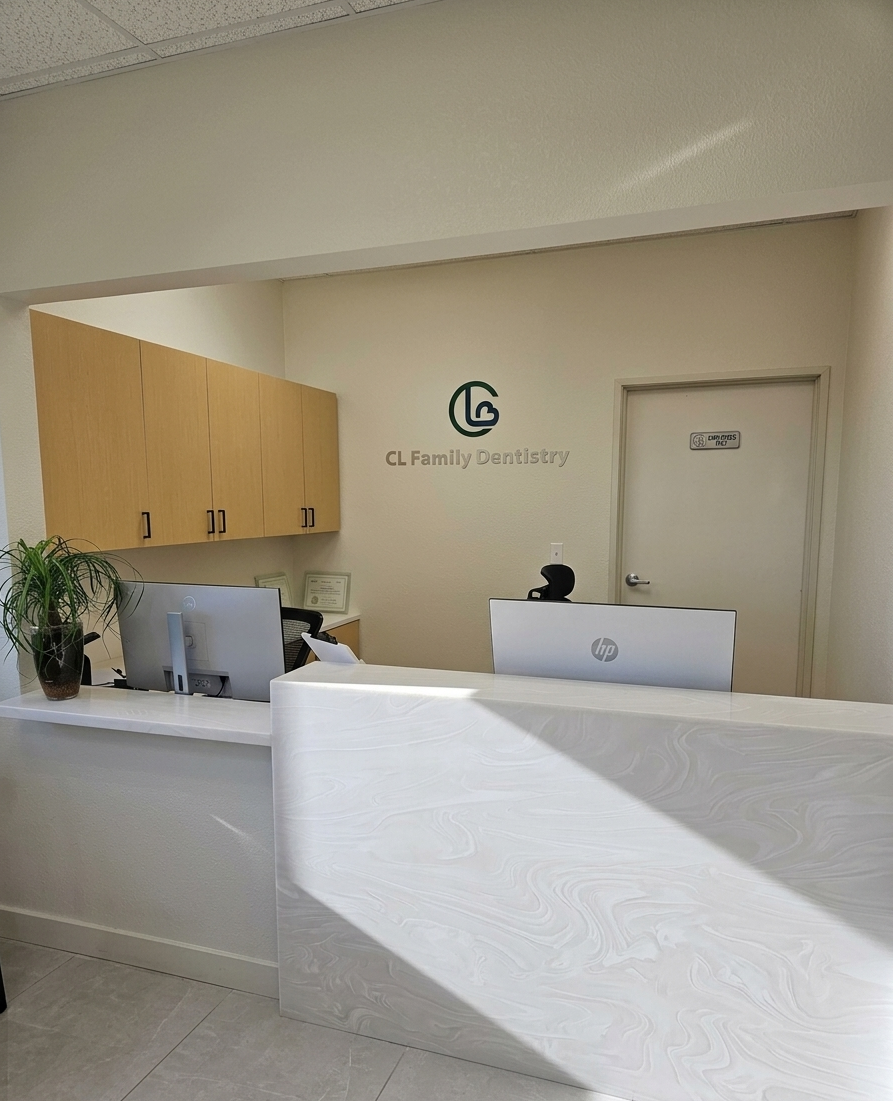 A portrait-oriented photograph captures a well-lit reception desk inside a dental office. Behind the counter are two computer monitors on a work surface, a potted plant, wooden cabinets, a logo on the back wall reading "CL Family Dentistry," and a door. The floor has light gray square tiles, and the ceiling is tiled with an acoustic texture.