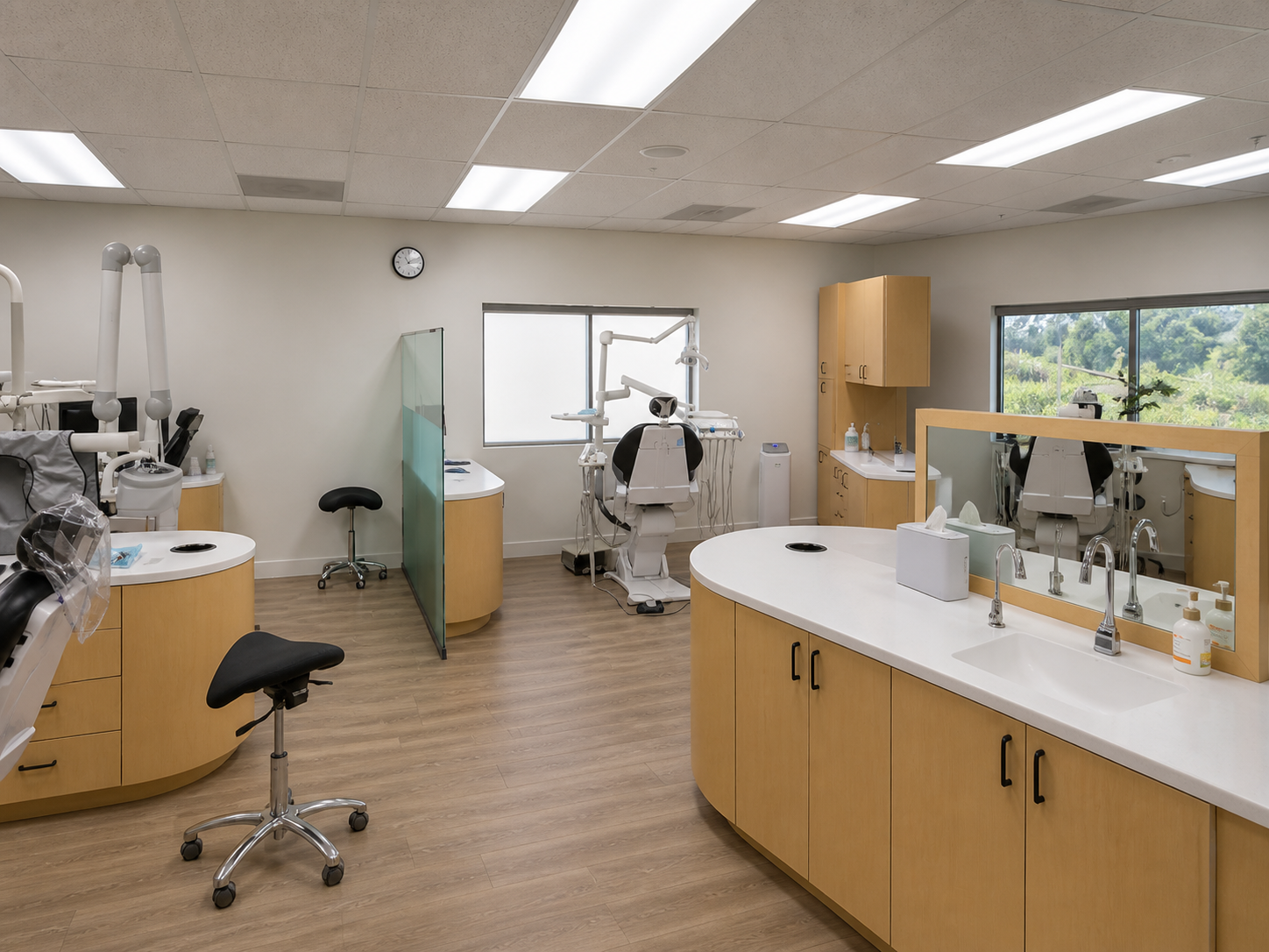 A spacious dental operatory with three treatment stations featuring golden cabinetry, white sinks, dental chairs, modern equipment, large windows overlooking trees, bright overhead lighting, and a clean wooden floor.