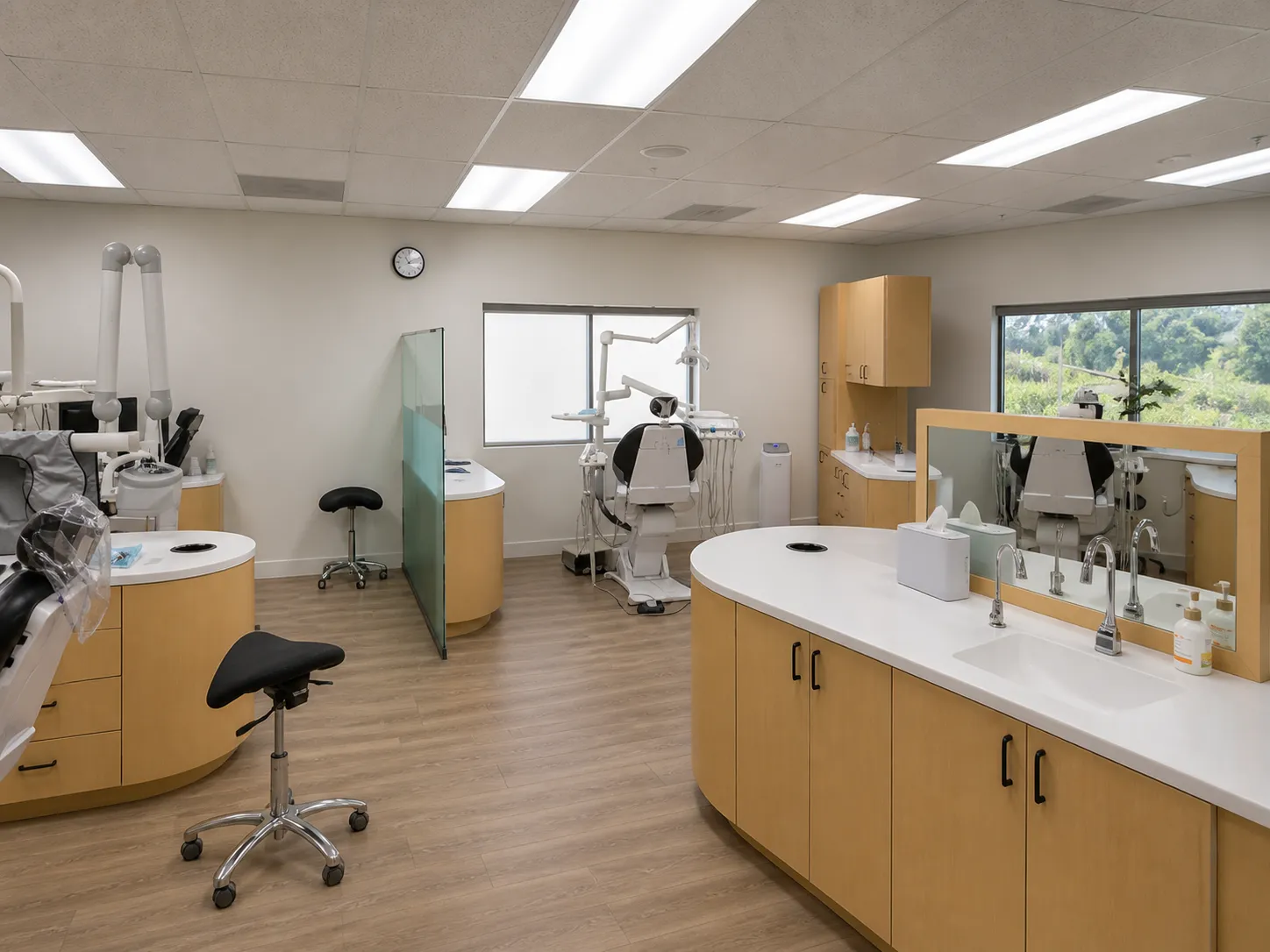 A spacious dental operatory with three treatment stations featuring golden cabinetry, white sinks, dental chairs, modern equipment, large windows overlooking trees, bright overhead lighting, and a clean wooden floor.