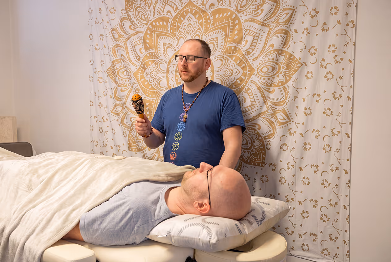 Therapist holding a rattle tool near a relaxed man lying on a massage table with a blanket and pillow.