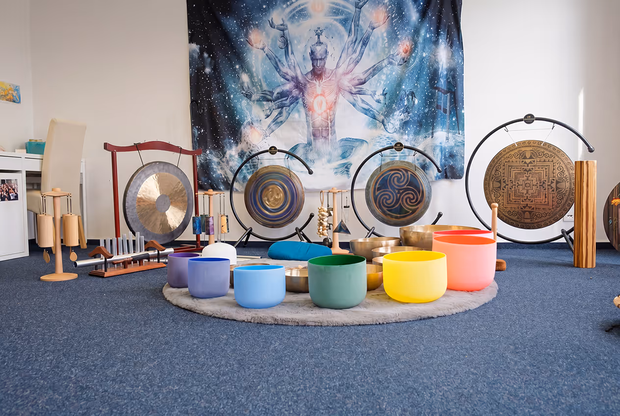 Room with various colorful singing bowls, gongs, and percussion instruments arranged on a round rug, with a mystical blue tapestry featuring a multi-armed figure hanging on the wall.