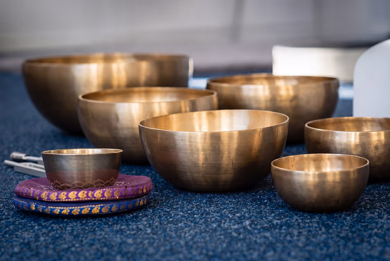 Set of brass singing bowls of various sizes arranged on a blue carpet with two patterned cushions beneath one small bowl.