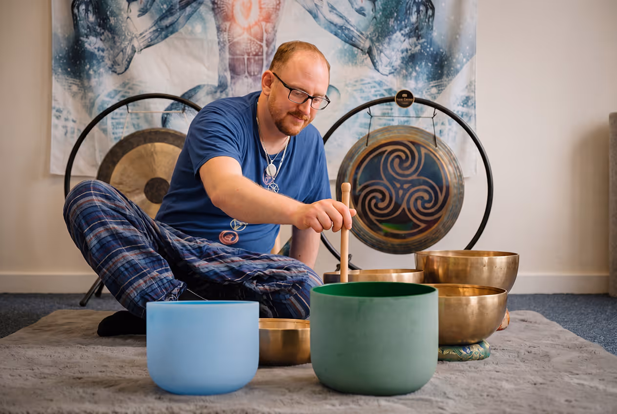Man sitting cross-legged on a carpet playing singing bowls with a mallet in a room decorated with gongs and a mystical wall hanging.