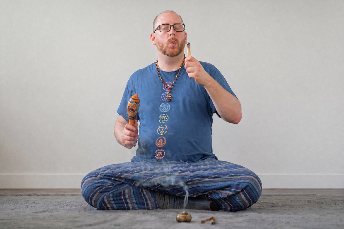 Smiling man with glasses sitting cross-legged on a yoga mat in a bright room with wooden accents.