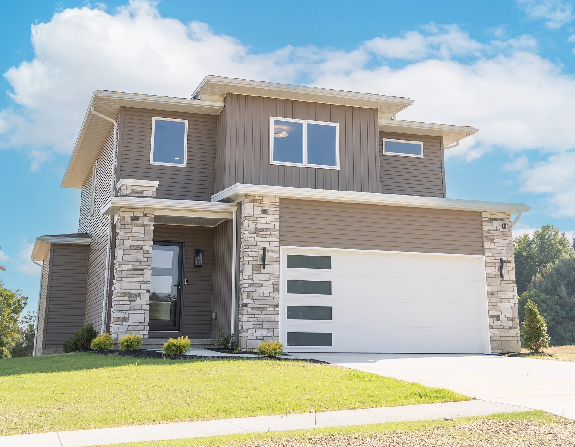 Modern two-story house with stone and brown siding, large garage, and green lawn under a blue sky.