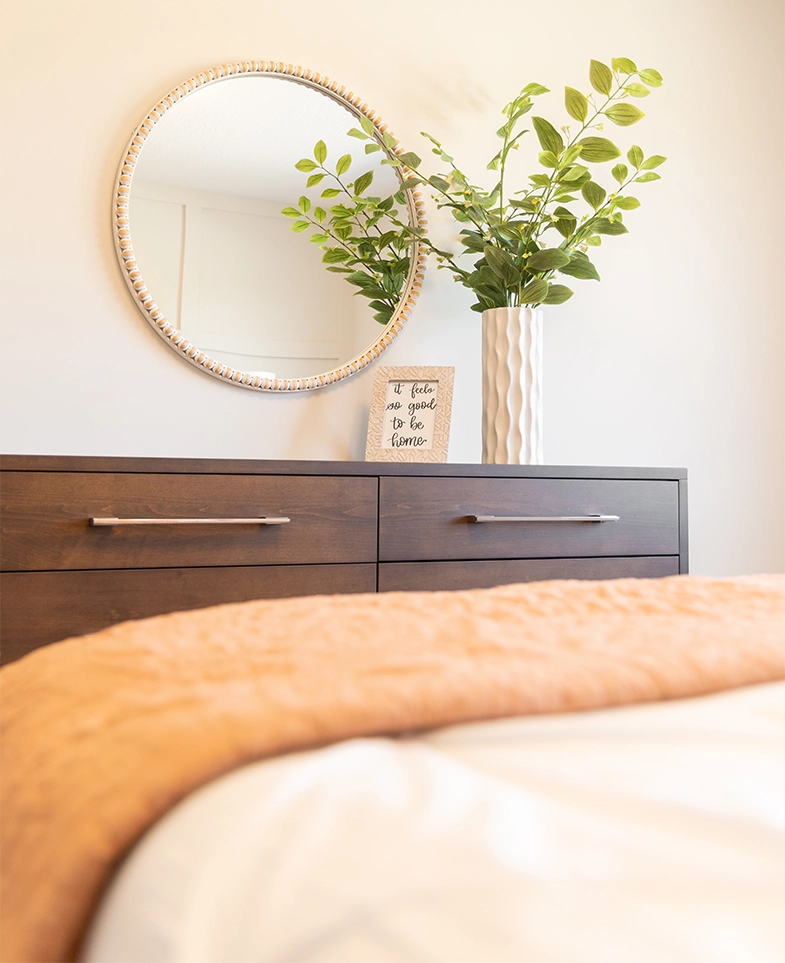Interior view of a bedroom showing a dark wooden dresser with silver handles, a round wall mirror with a decorative frame, a tall white textured vase containing green leafy branches, and a framed sign reading 'It feels so good to be home.' The foreground shows a blurred beige and white bedspread.