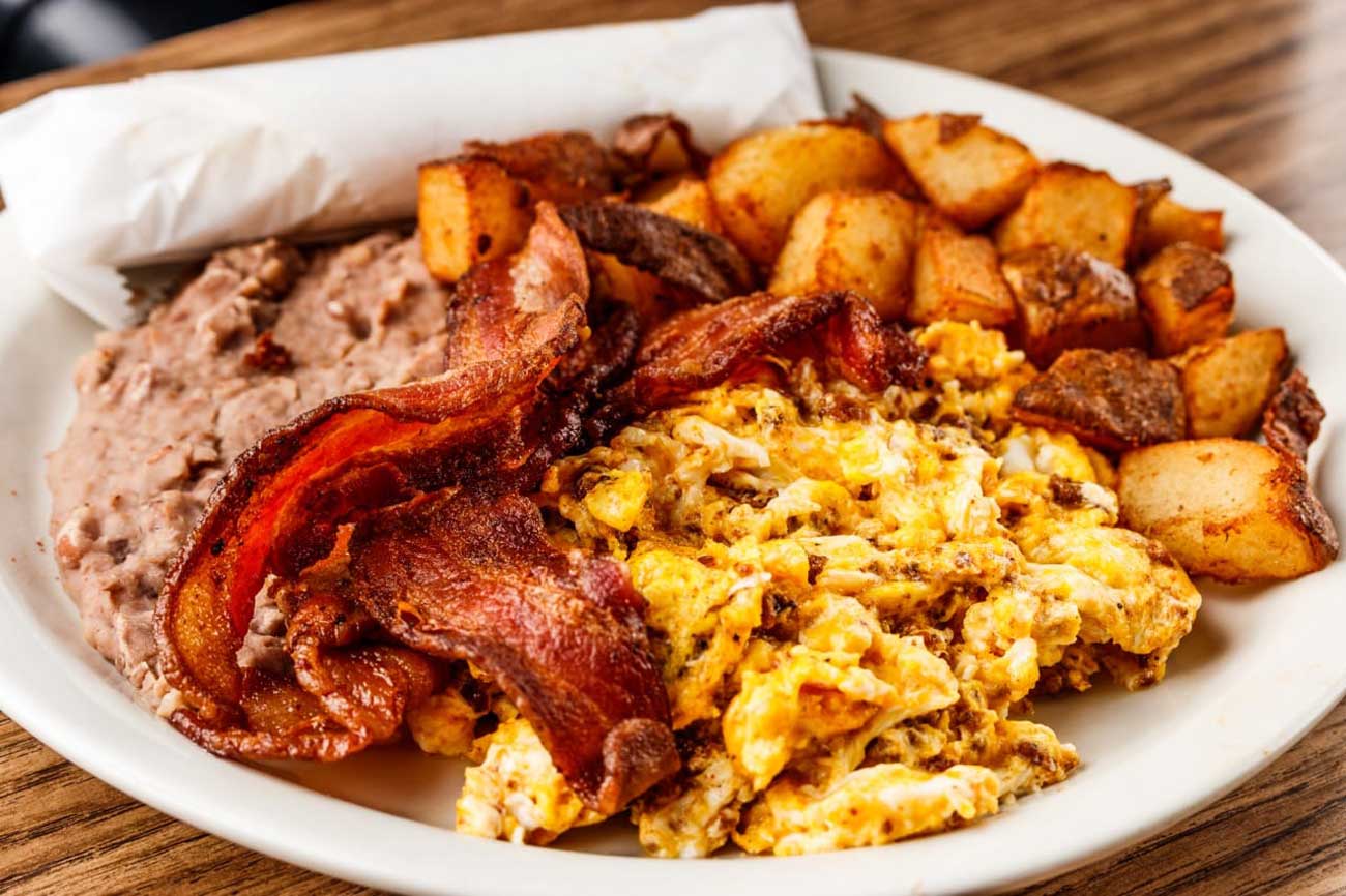 Plate with crispy bacon, scrambled eggs, seasoned breakfast potatoes, refried beans, and a wrapped flour tortilla.