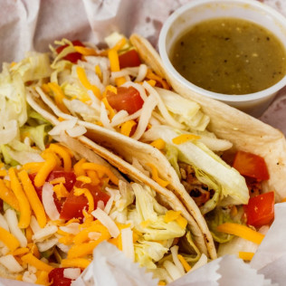 Two soft tacos filled with shredded cheese, lettuce, and diced tomatoes, served with a side of green salsa in a plastic container.