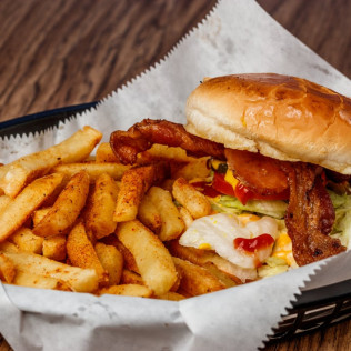 Bacon cheeseburger with lettuce, tomato, and ketchup served with seasoned French fries in a basket on parchment paper.