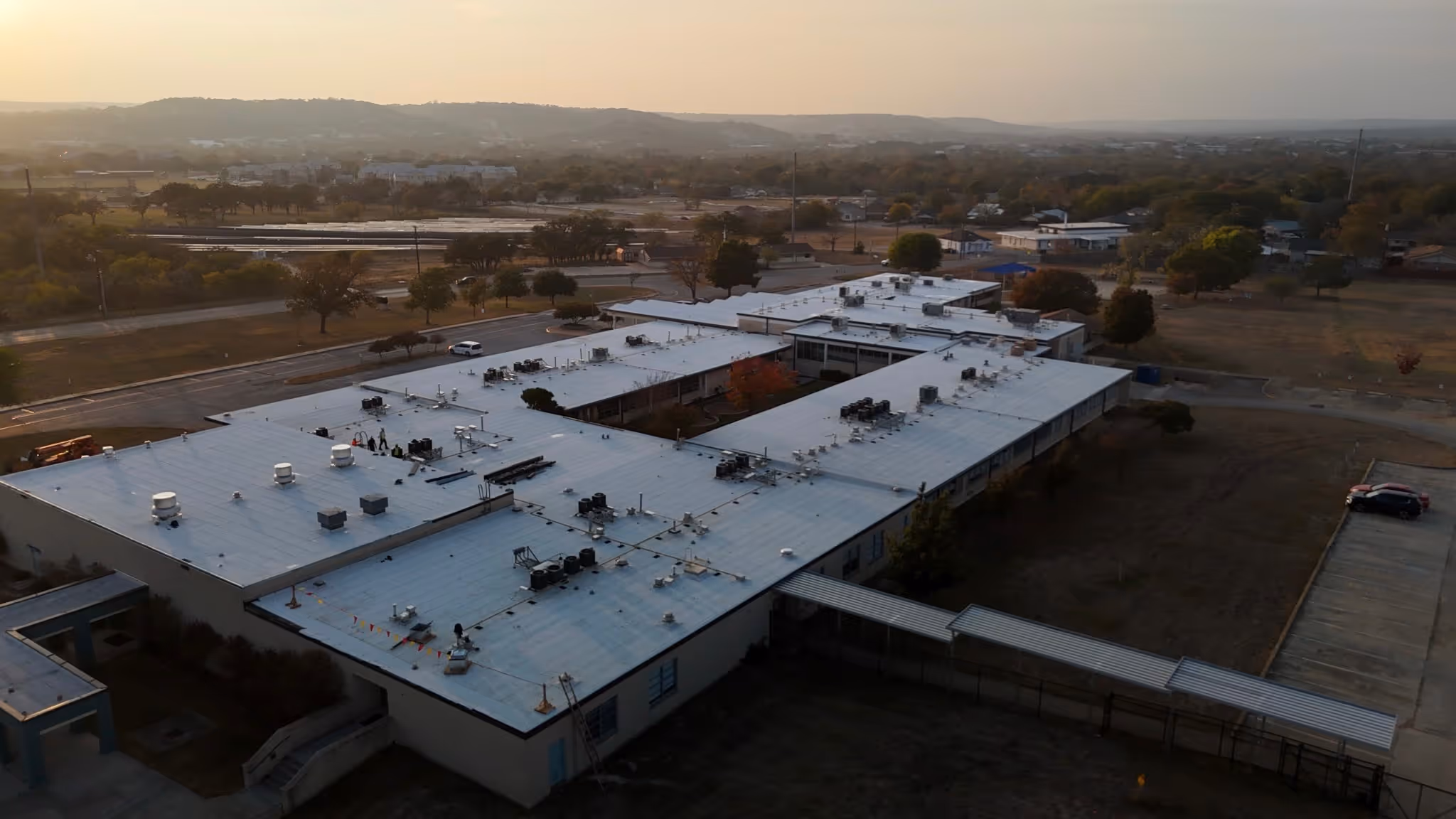 Aerial view of a large, single-story building with a flat roof and courtyard in a suburban area at sunset.