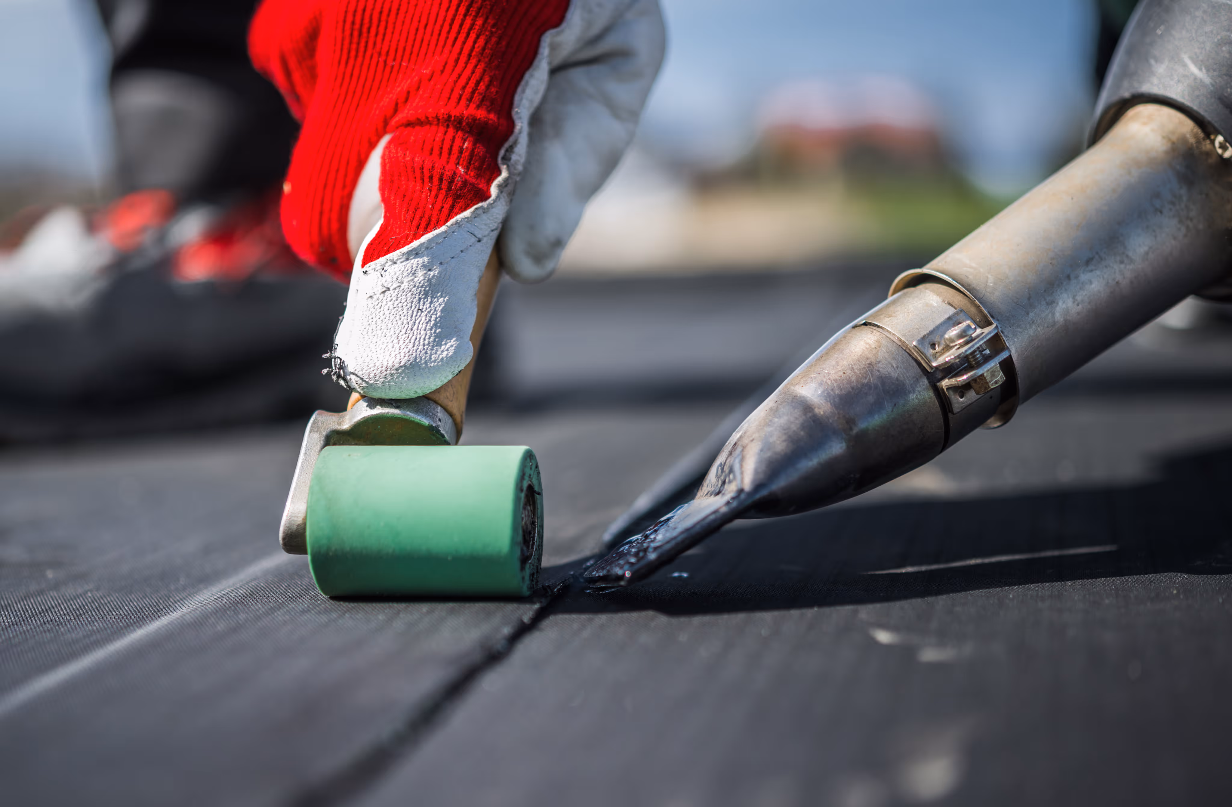 Hand with red and white glove pressing a green roller to seal black roofing material with heat.