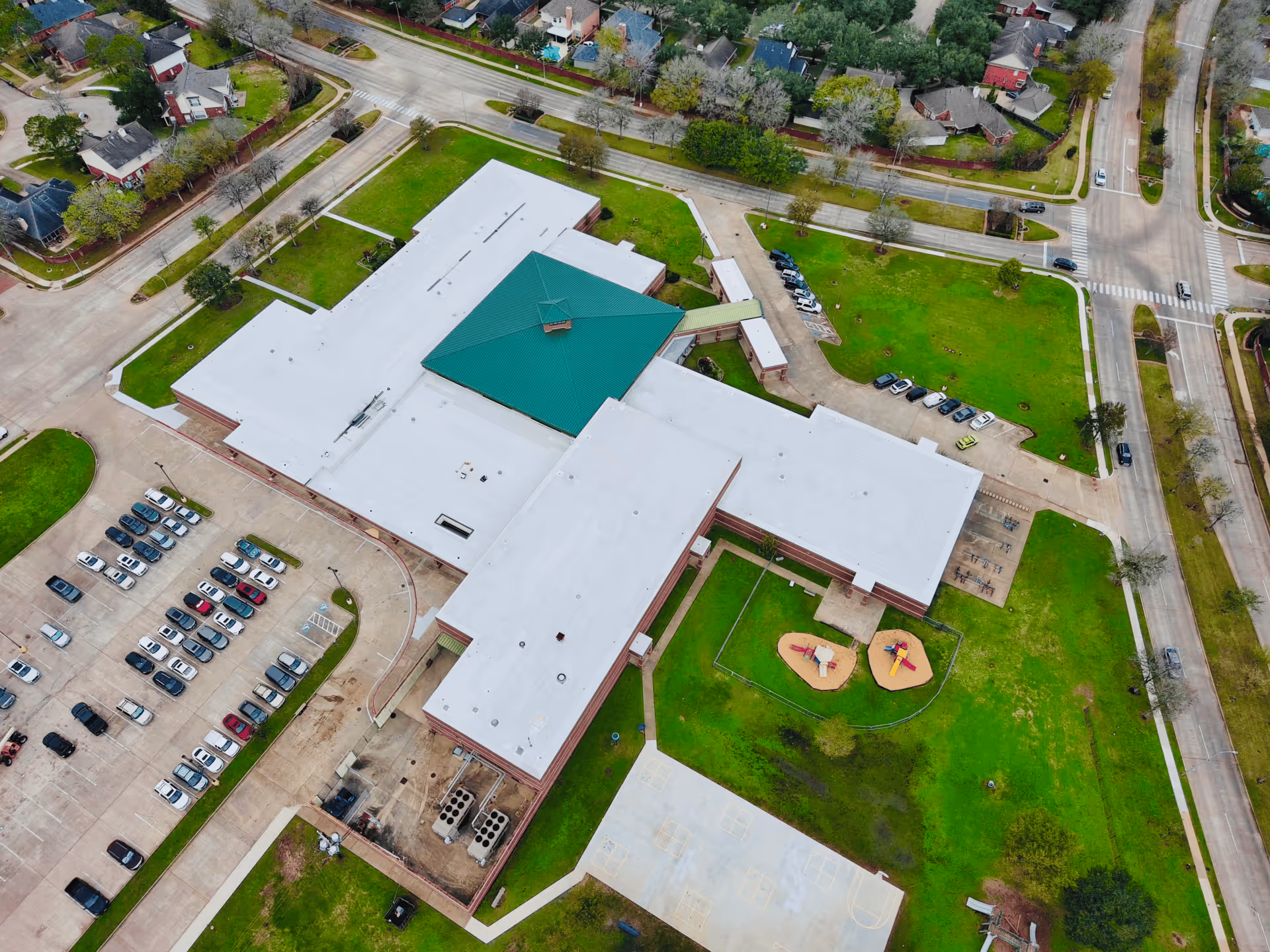 Aerial view of a large building with a green roof section, adjacent parking lot, playground, and surrounding green spaces.
