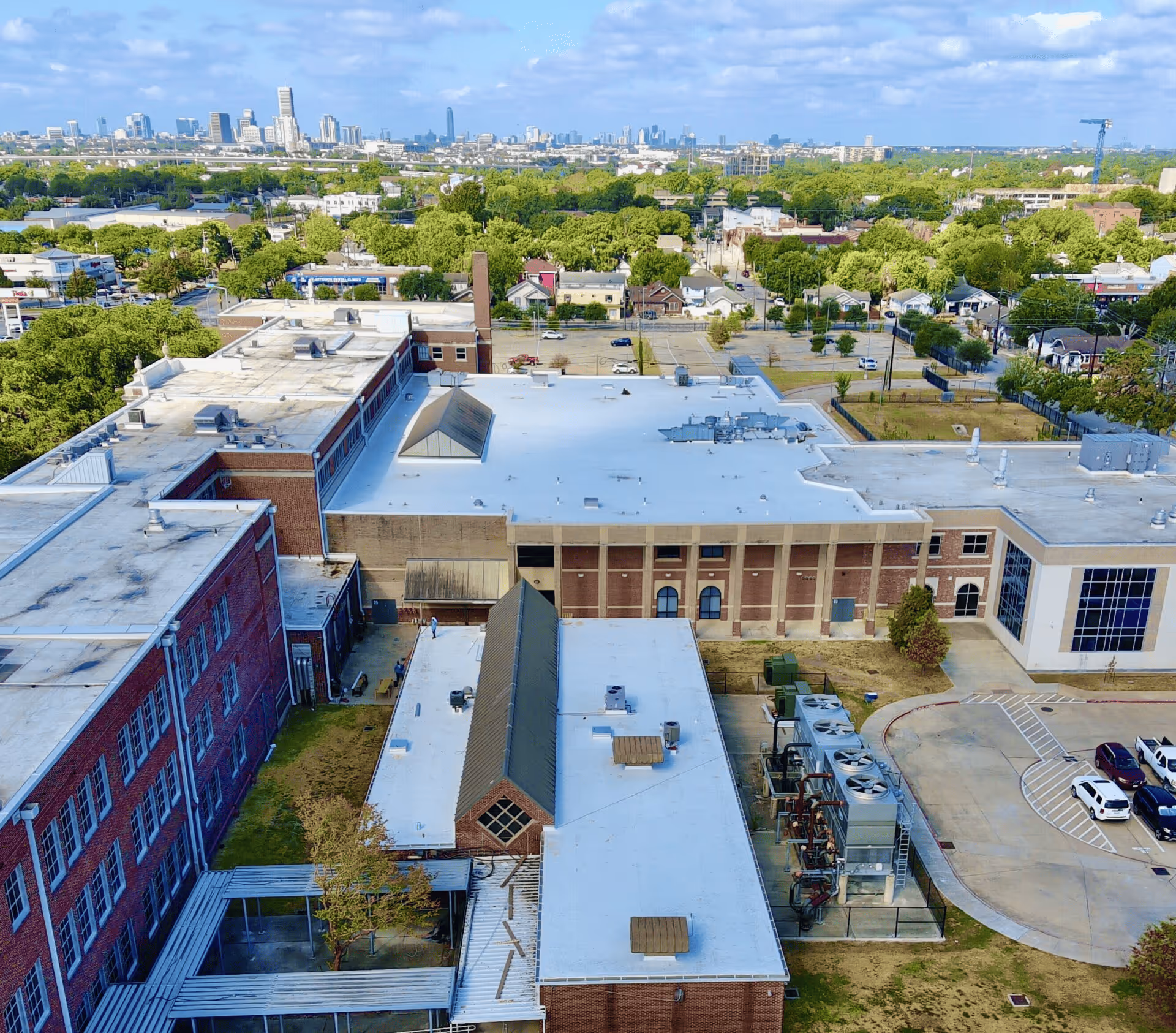 Aerial view of brick buildings with white rooftops amid green trees and city skyline in the background under a blue sky.