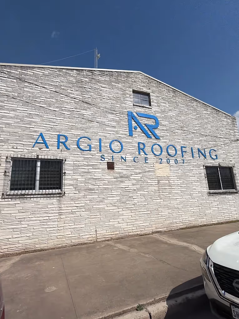 White brick building with blue sign reading AR Argio Roofing Since 2007 under clear blue sky.