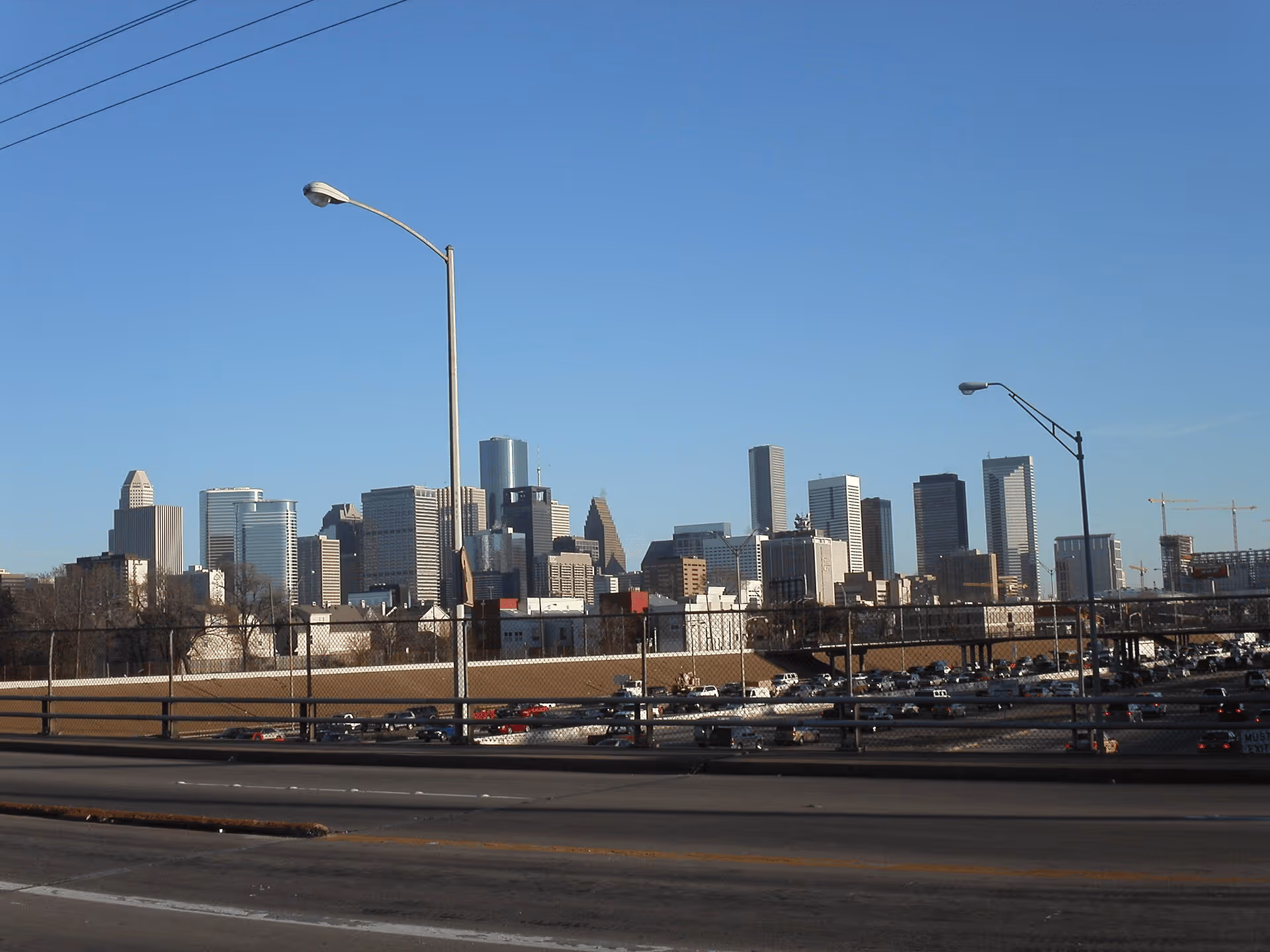 City skyline with modern skyscrapers under a clear blue sky, viewed from a highway with street lamps and traffic below.