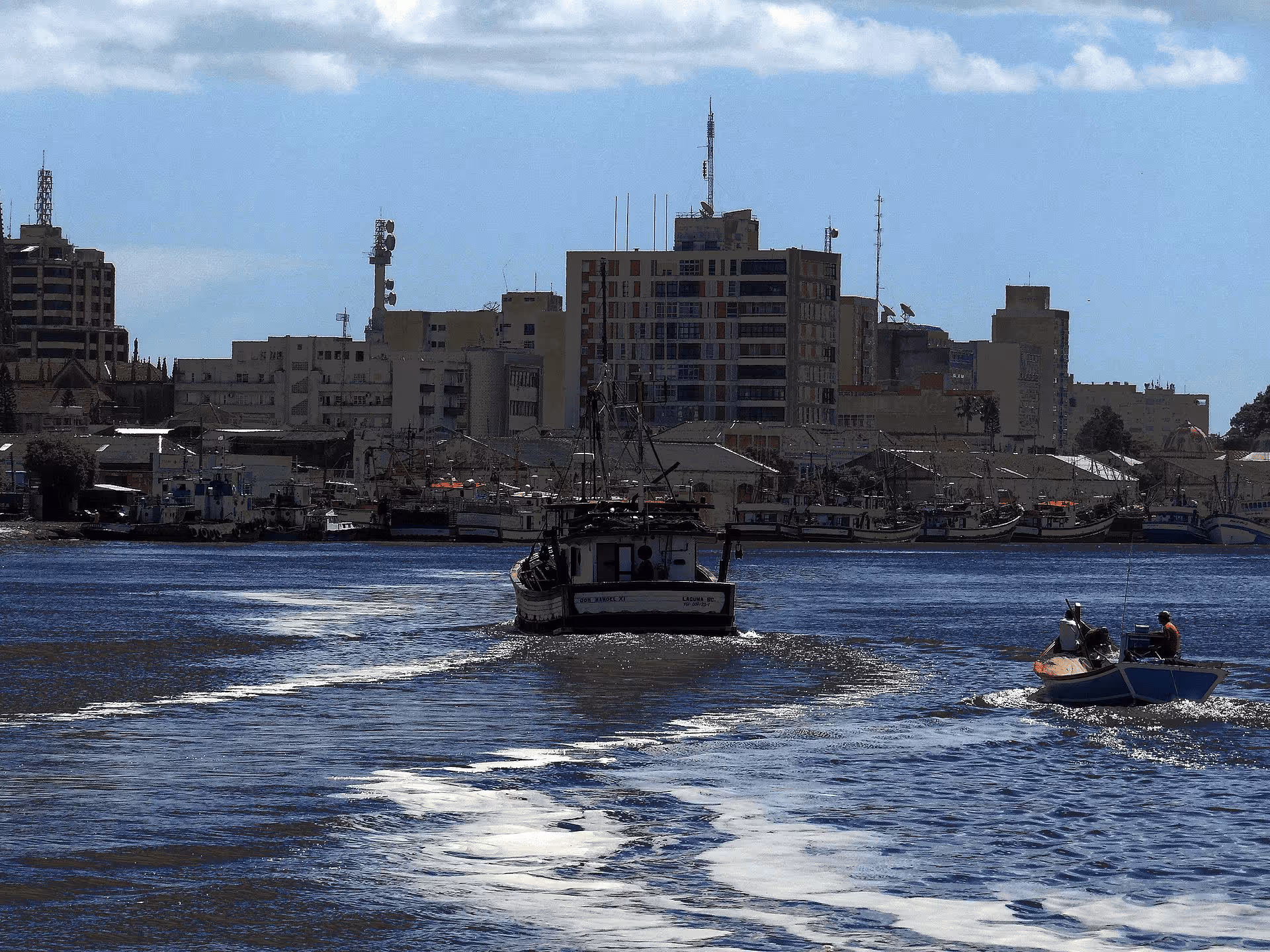 Fishing boats on water with city buildings in the background under a blue sky with clouds.