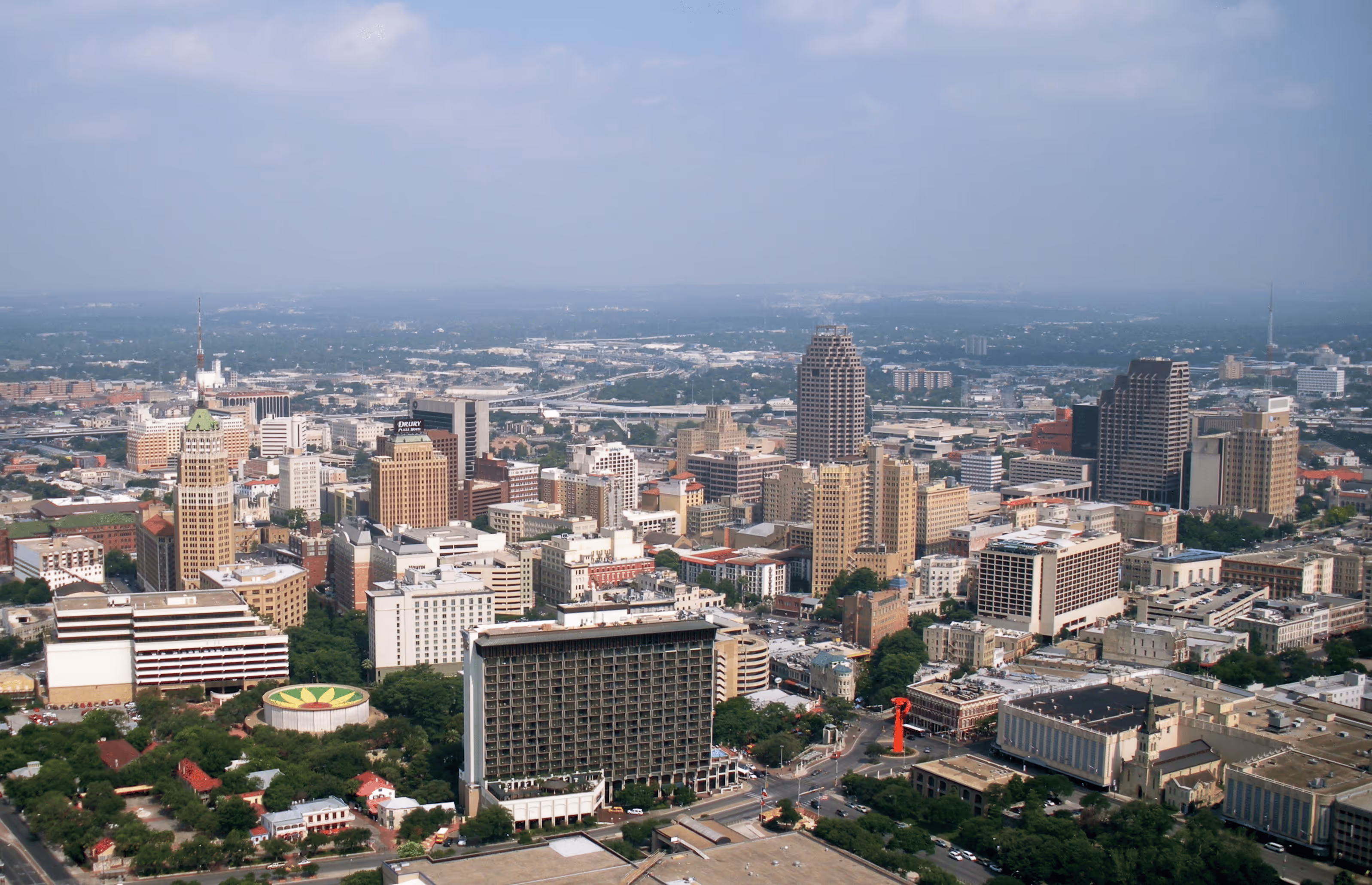 Aerial view of a city skyline with various high-rise buildings and green trees under a partly cloudy sky.