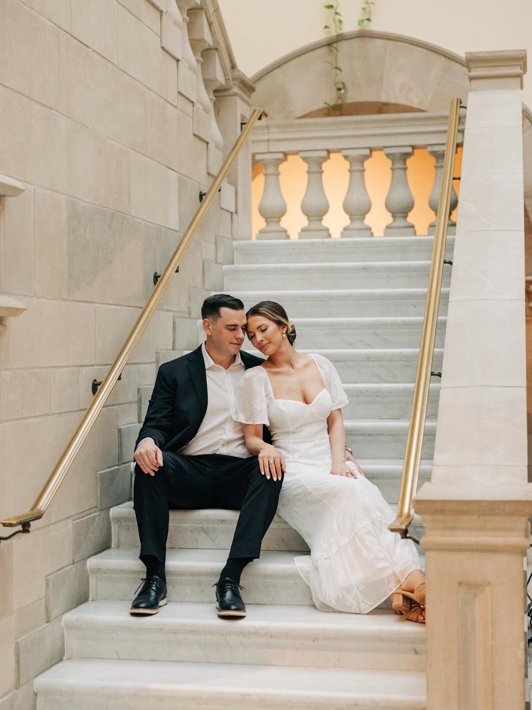 A couple sitting close together on marble stairs inside a building with beige stone walls and golden handrails, the woman wearing a white dress and the man in a dark suit with a white shirt.