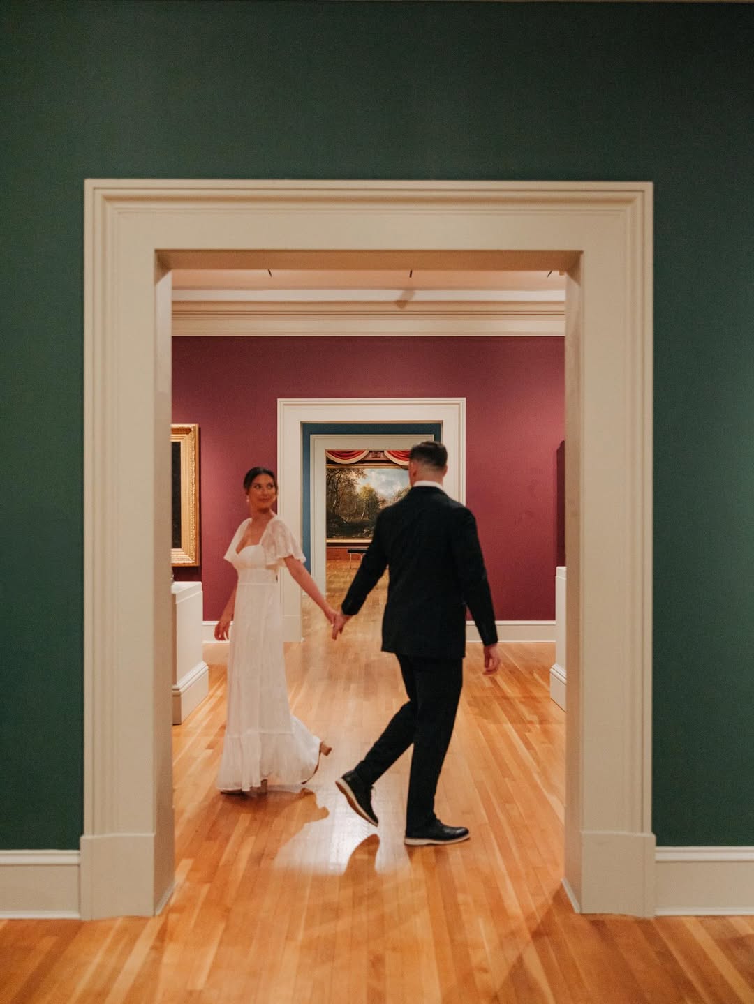 A couple holding hands walking through a doorway in an art gallery with green and dark red walls and wooden floors. The woman wears a white dress and looks back while the man in a black suit leads forward.