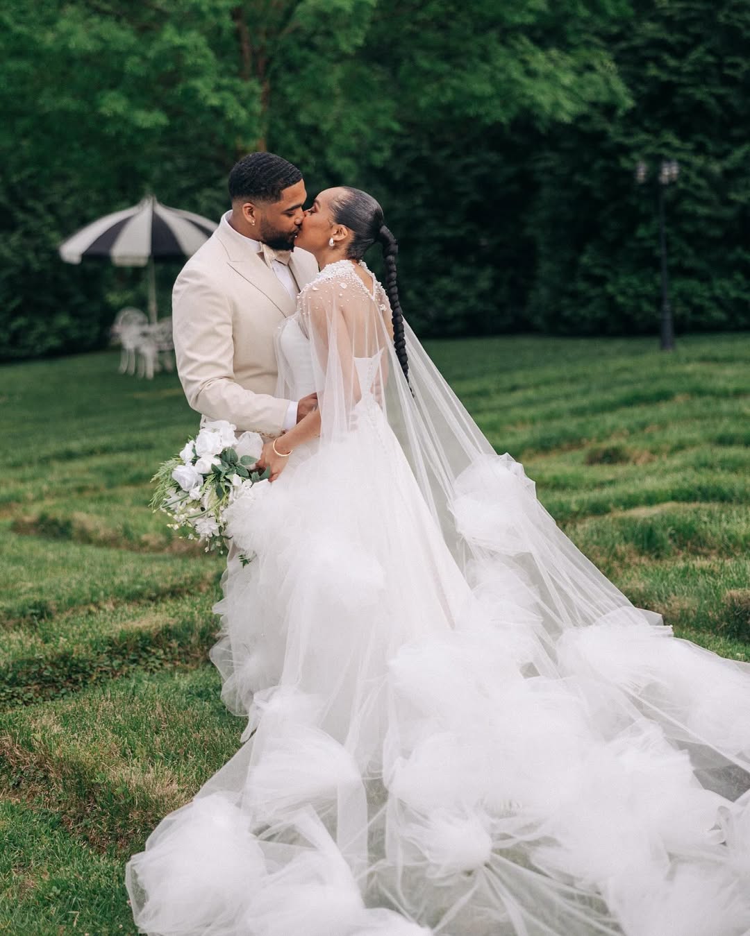 Bride and groom sharing a kiss outdoors on grass, with the bride wearing a long white wedding dress with a flowing veil and holding a bouquet of white flowers, and the groom in a light-colored suit.