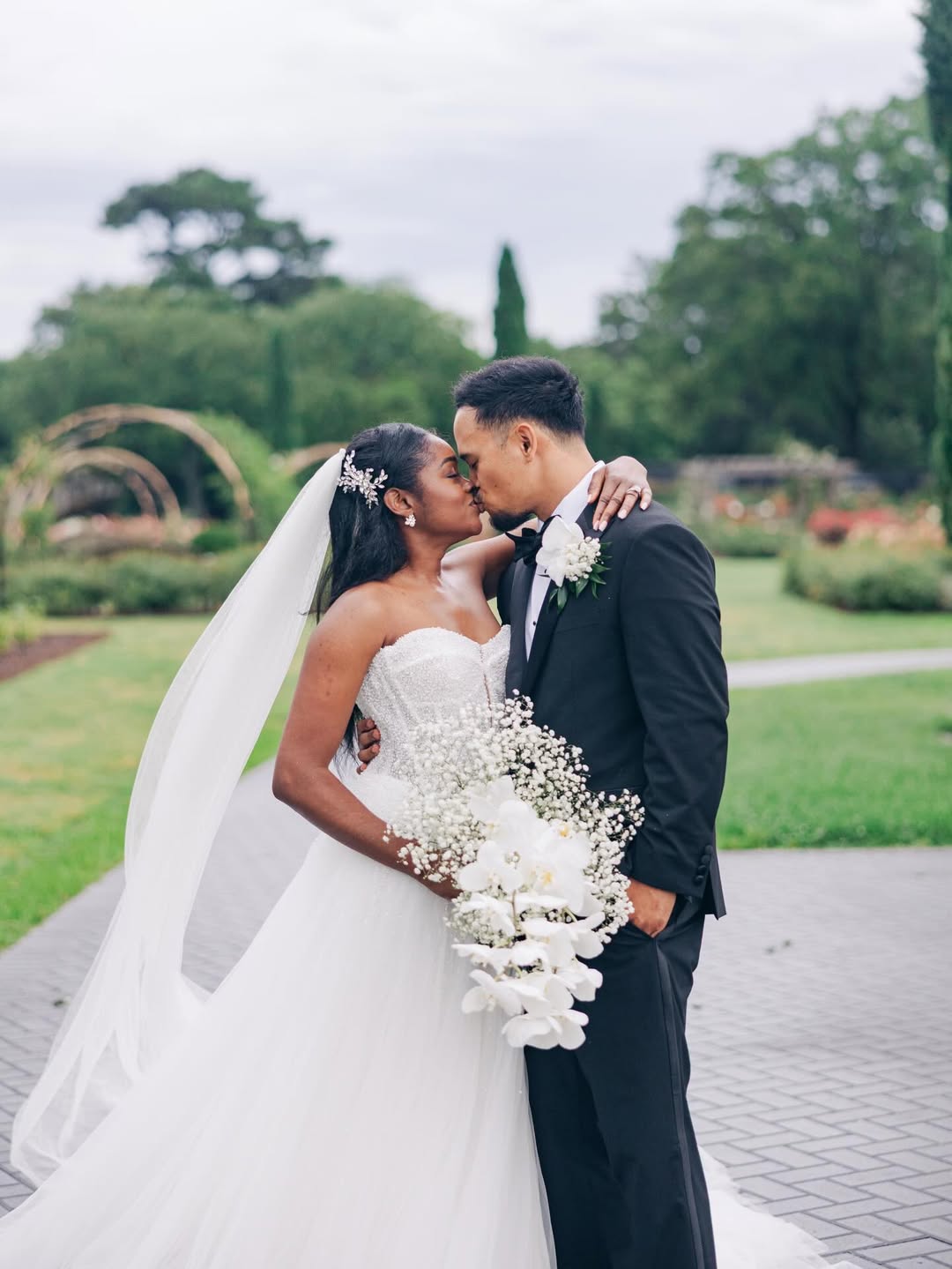 Bride and groom kissing outdoors on a paved pathway, with the bride wearing a strapless white gown and veil holding a cascading bouquet of white flowers, and the groom in a black tuxedo with a white boutonniere, surrounded by greenery and trees.