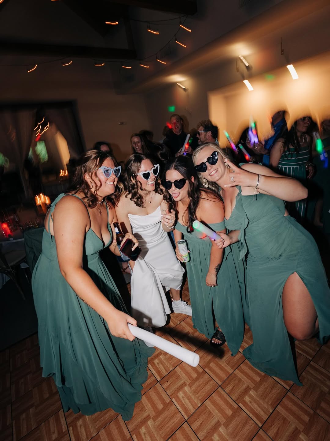 Four women in dresses and heart-shaped sunglasses pose happily on a dance floor at a party, holding glow sticks and drinks.