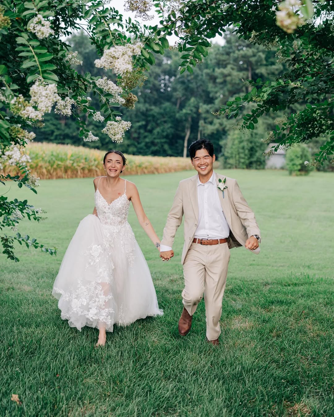 A joyful bride in a white floral lace wedding dress and a groom in a beige suit holding hands and walking barefoot on green grass under leafy branches outdoors.