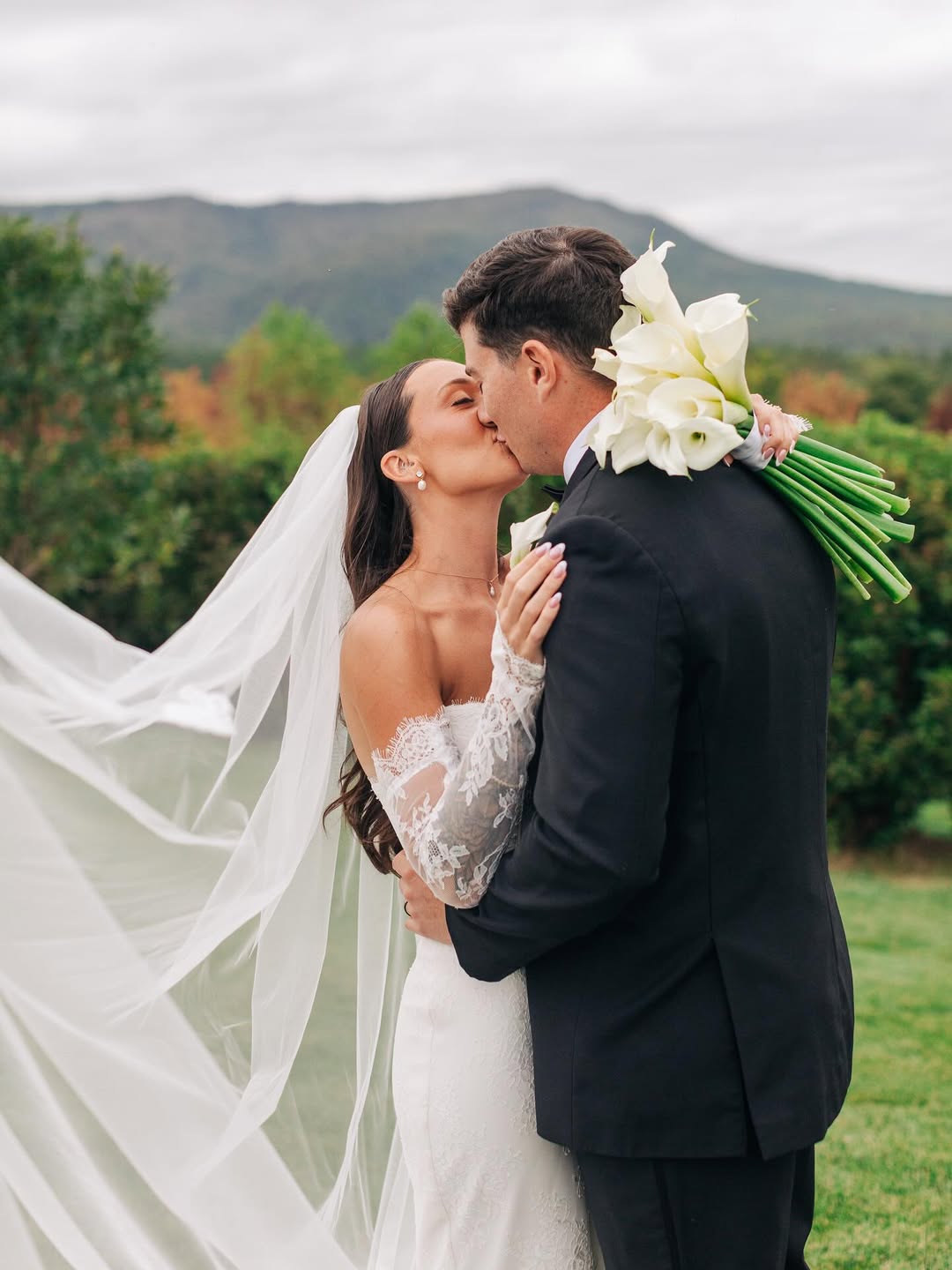 Bride and groom kissing outdoors with the bride wearing a white lace wedding dress and veil, and the groom in a black suit holding a bouquet of white calla lilies, with a background of green bushes and distant hills.