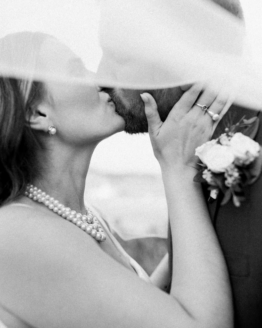 Black and white close-up photo of a couple kissing, with the woman holding the man's face gently, wearing a pearl necklace and earrings, and the man wearing a boutonniere.
