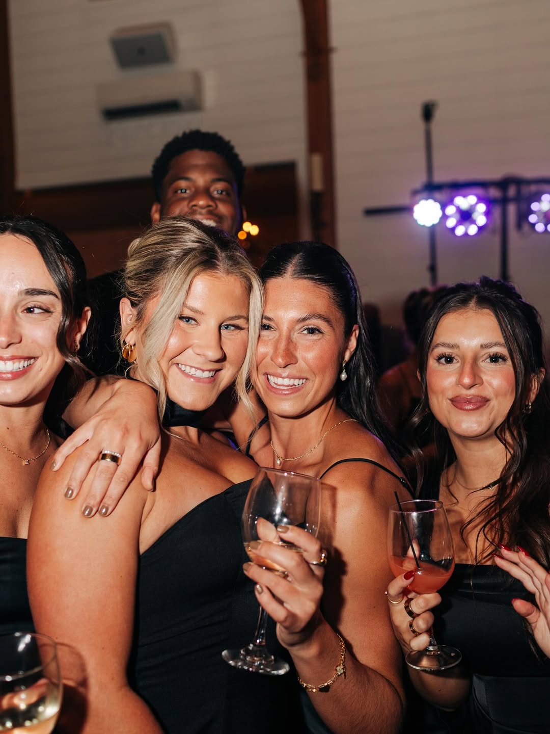 Group of four women and one man smiling at a party, three women hold wine glasses with drinks, dressed in black outfits, with purple party lights in the background.