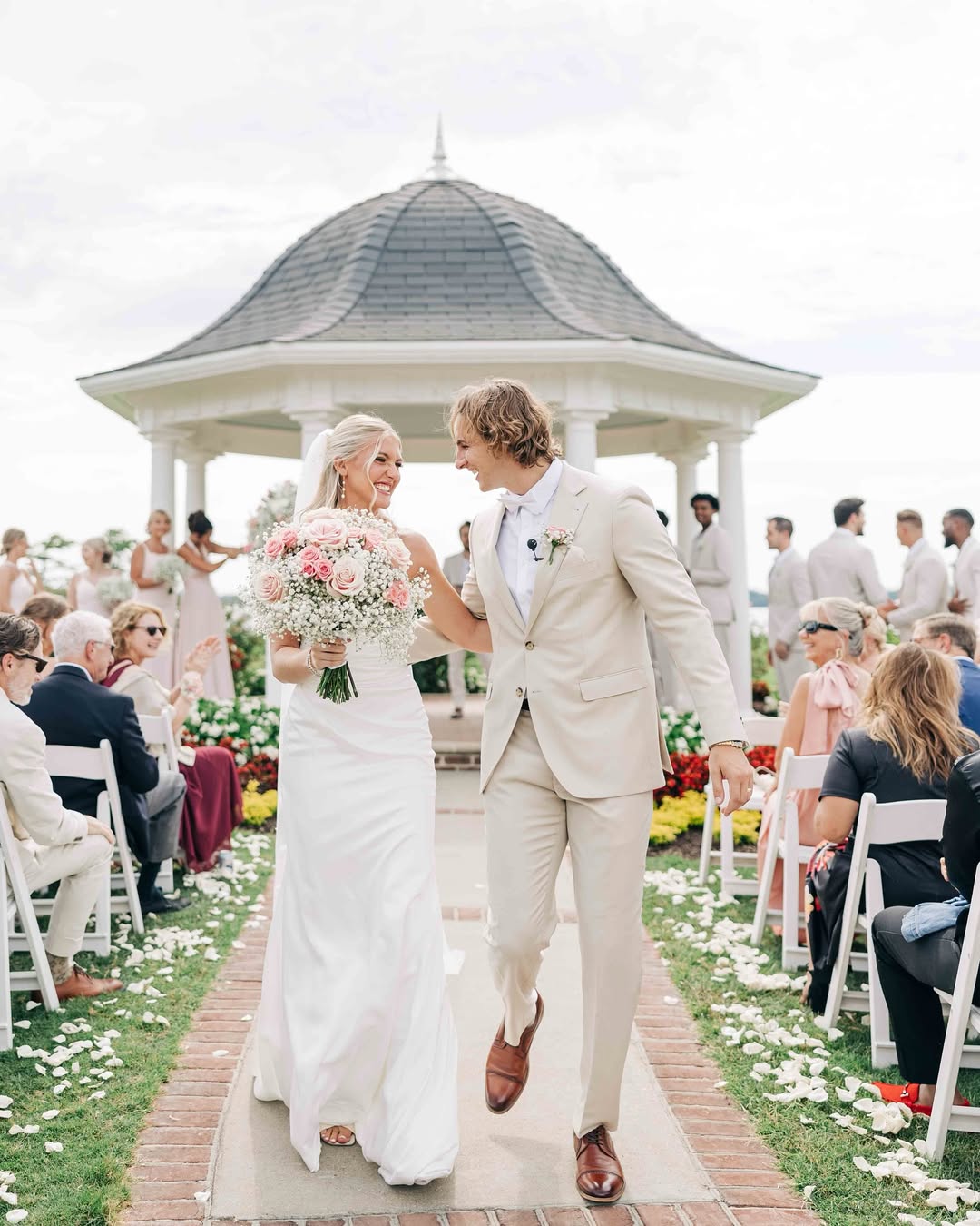 Bride and groom walking down an outdoor aisle smiling at each other, with the bride holding a bouquet of pink and white roses and baby's breath, guests seated on either side, and a white gazebo in the background.