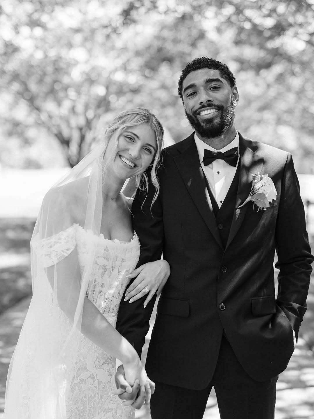 Smiling bride in lace wedding dress and veil holding hands with groom in black tuxedo and bow tie, outdoors with blurred trees in background.