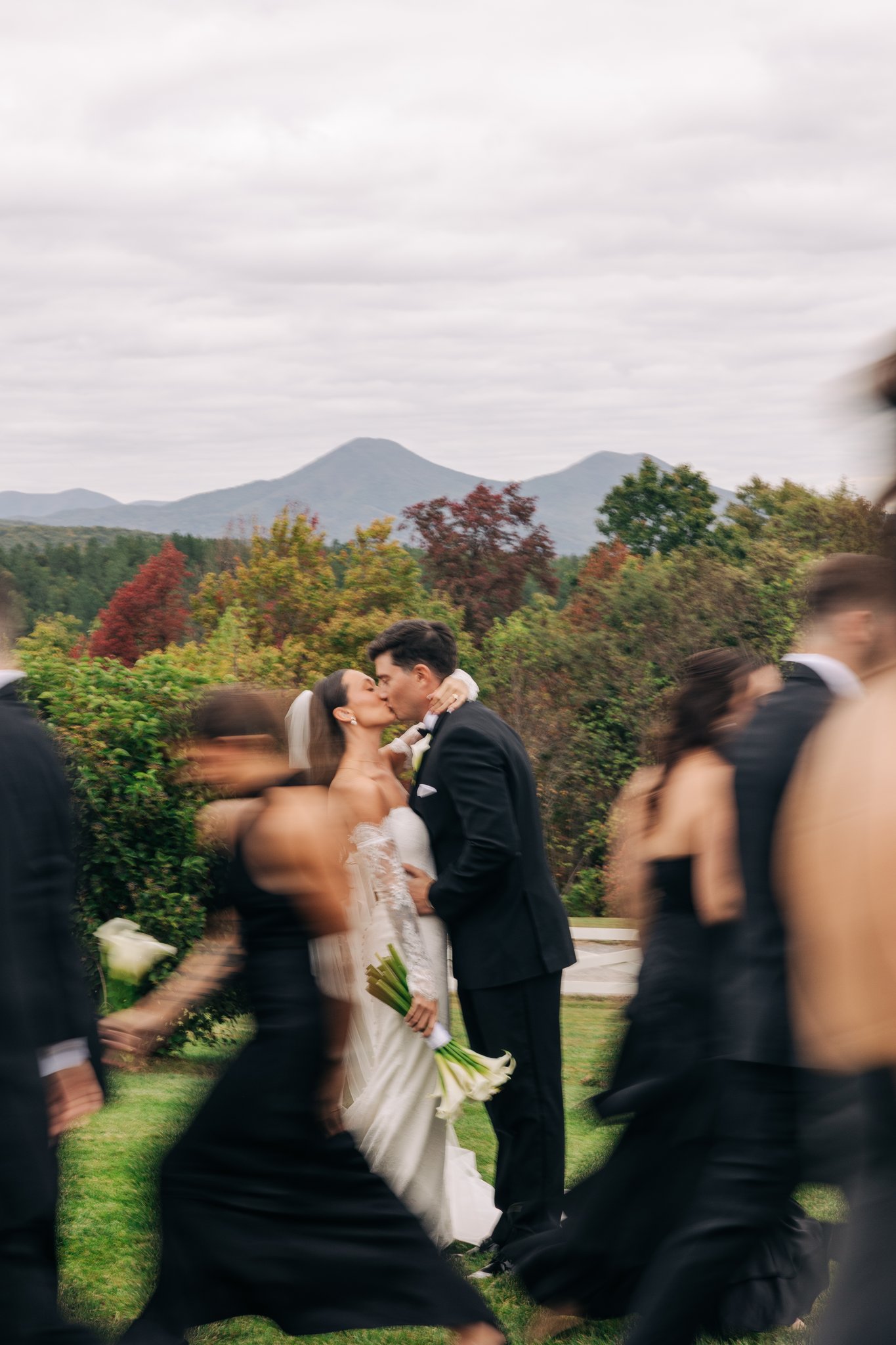 Bride and groom kissing outdoors on grass with blurred wedding party members in black dresses and suits walking around them, with trees and mountains in the background under a cloudy sky.